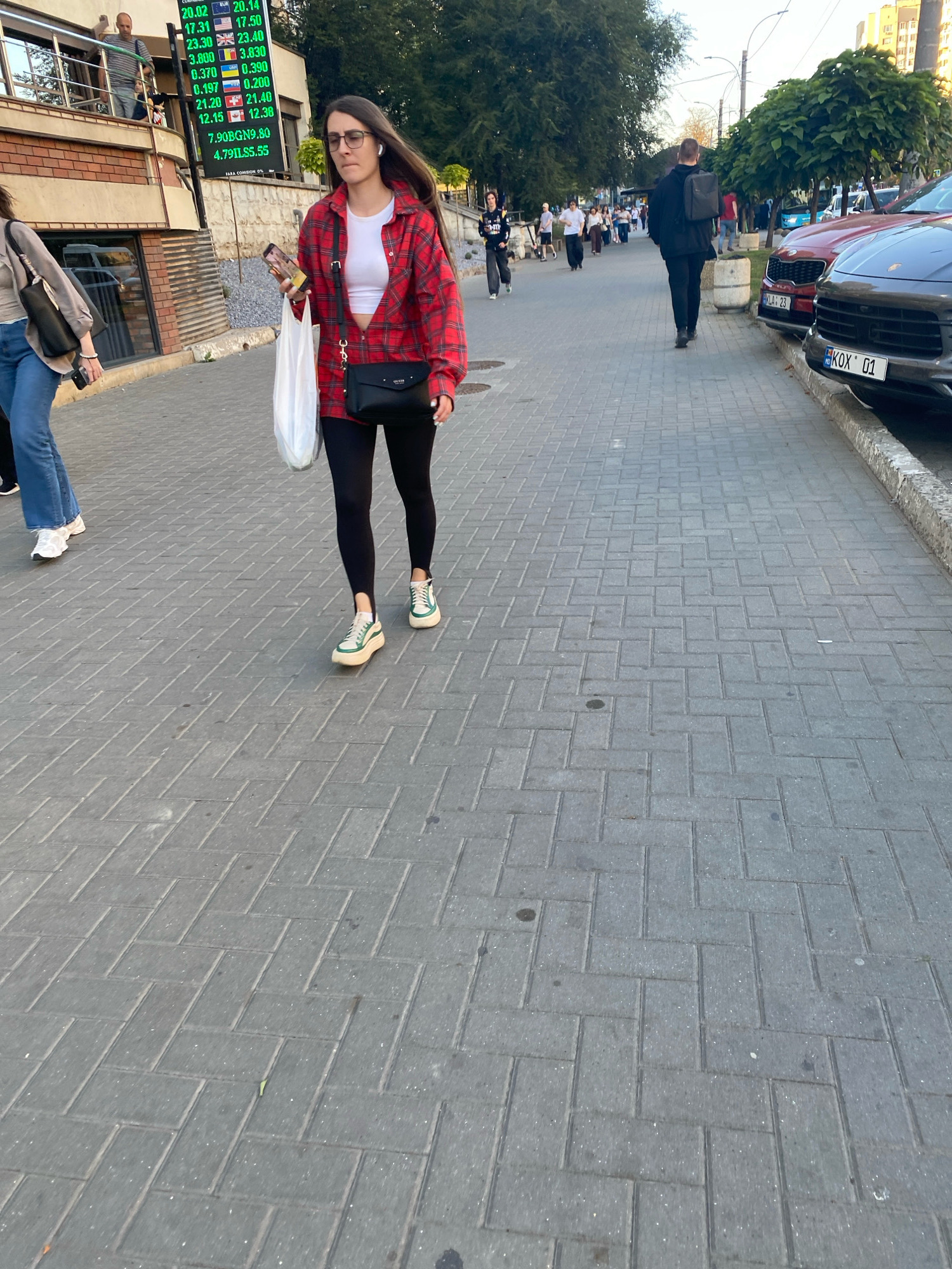 A young woman with long brown hair, wearing glasses and wireless earbuds, walks on a paved sidewalk in Chișinău, Moldova. She is dressed in a red and black plaid shirt over a white crop top, black leggings, and green and white platform sneakers. She carries a white plastic bag in her left hand and a black crossbody bag over her right shoulder. The background shows a street with parked cars, buildings, trees, and other pedestrians walking in the distance. A digital display board showing currency exchange rates is visible on a building to the left. The license plate on the dark gray car in the foreground reads "KDX 01".