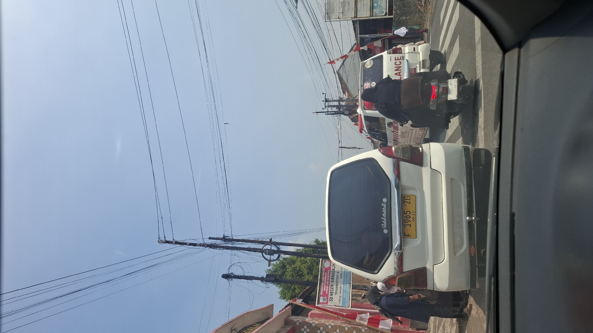 In a street scene in Sirnagalih, Indonesia, an overcast sky looms above a traffic intersection. In the foreground, the rear of a white SUV is visible, its license plate reading "F 1965 ZG". To the left, two individuals, one wearing a dark hijab and the other partially visible, stand near a building with red and white trim. Adjacent to the SUV, an ambulance, marked with "LANCE", is stopped in traffic. Behind the ambulance, a scooter with two occupants, both wearing dark headscarves, is positioned at the crosswalk. The environment includes power lines, utility poles, and roadside buildings. A sign identifying "SD NEGERI SIRNAGALIKH" is mounted on a pole.