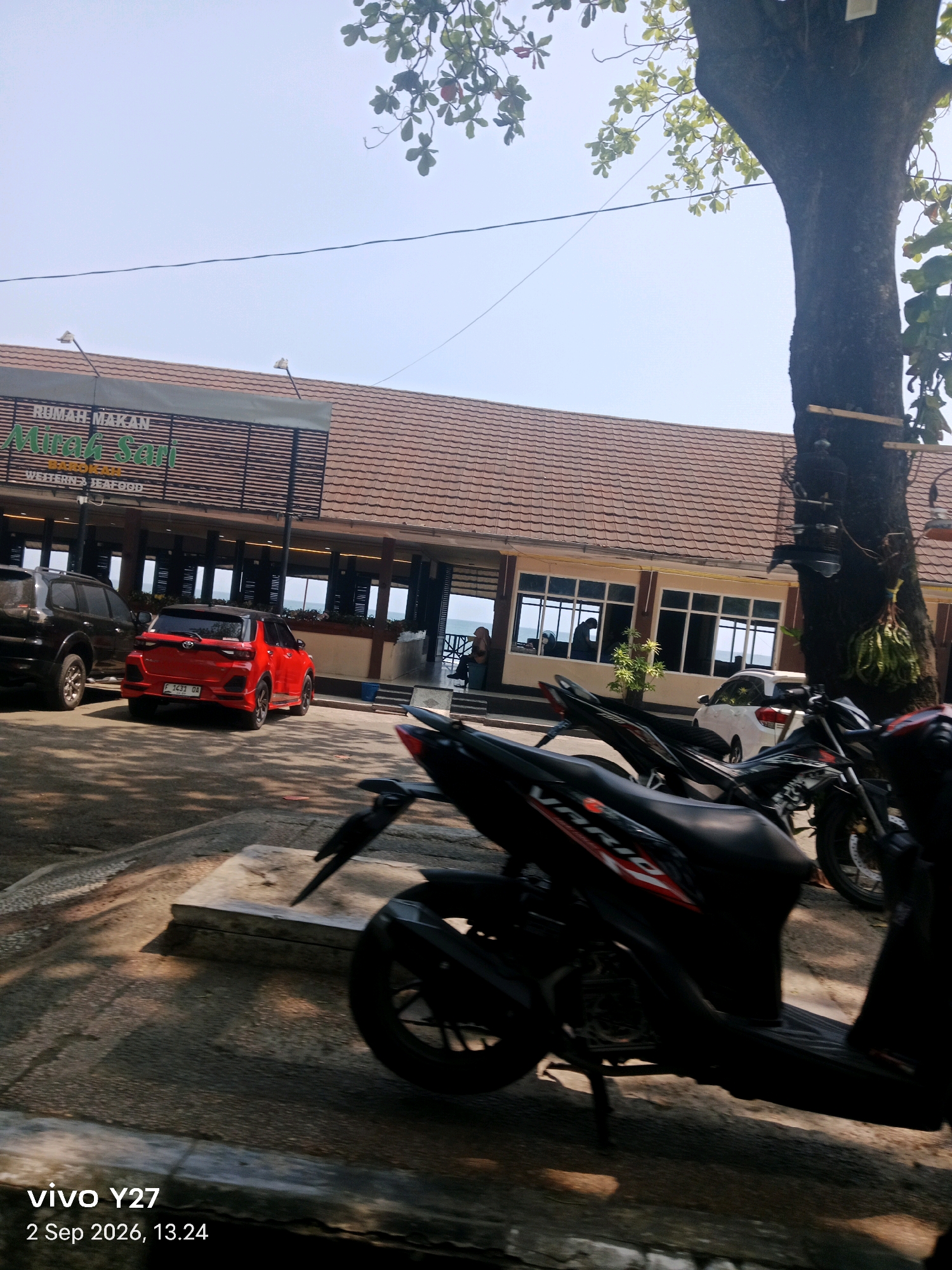 The image depicts an outdoor scene in front of a building identified by a sign as "RUMAH MAKAN" (Restaurant) with the name "Mirah Sari" prominently displayed, suggesting it is located in Citepus, Indonesia. The restaurant has a reddish-brown tiled roof and a beige facade with large windows. In the foreground, several motorcycles are parked, with one black scooter in the immediate foreground. To the left of the motorcycles, a red SUV is parked. Further to the left, a black SUV is visible. Beyond the parked vehicles, the building appears to be situated near a body of water, possibly the sea, as suggested by the horizon visible through the windows. The sky is clear and bright blue. A large tree with green leaves stands to the right of the frame, partially obscuring the building. The ground is paved. The timestamp "vivo Y27 2 Sep 2026, 13.24" is present in the bottom left corner.