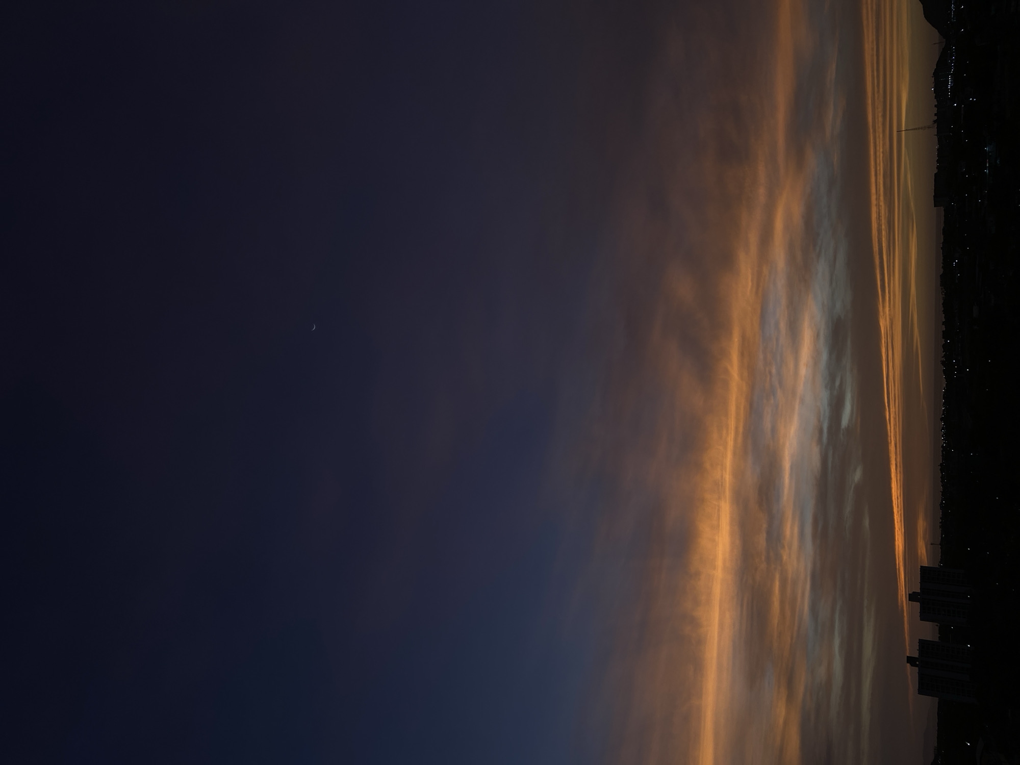 The image captures a serene twilight sky over Juan Griego, Venezuela, featuring a dramatic interplay of dark blues and warm, golden-orange cloud formations. The sky dominates the frame, transitioning from a very dark, deep blue-purple in the upper portion to vibrant hues of orange, gold, and lighter greyish-white closer to the horizon. These warm colors are reflected on streaky, elongated clouds, creating distinct bands of light that stretch across the sky, suggesting either a powerful sunset or sunrise.
A delicate, slender crescent moon is faintly visible in the darker, upper-left section of the sky. Below the colorful sky, a dark, silhouetted horizon line stretches across the bottom of the image. This silhouette reveals the dark shapes of distant hills or mountains on the right side and the outlines of several multi-story buildings on the left, indicative of an urban or semi-urban landscape. Small, scattered lights are visible along the horizon line, twinkling faintly against the fading light. The overall atmosphere is tranquil and visually striking, highlighting the beauty of nature's color palette at dusk or dawn. No text is visible in the image.