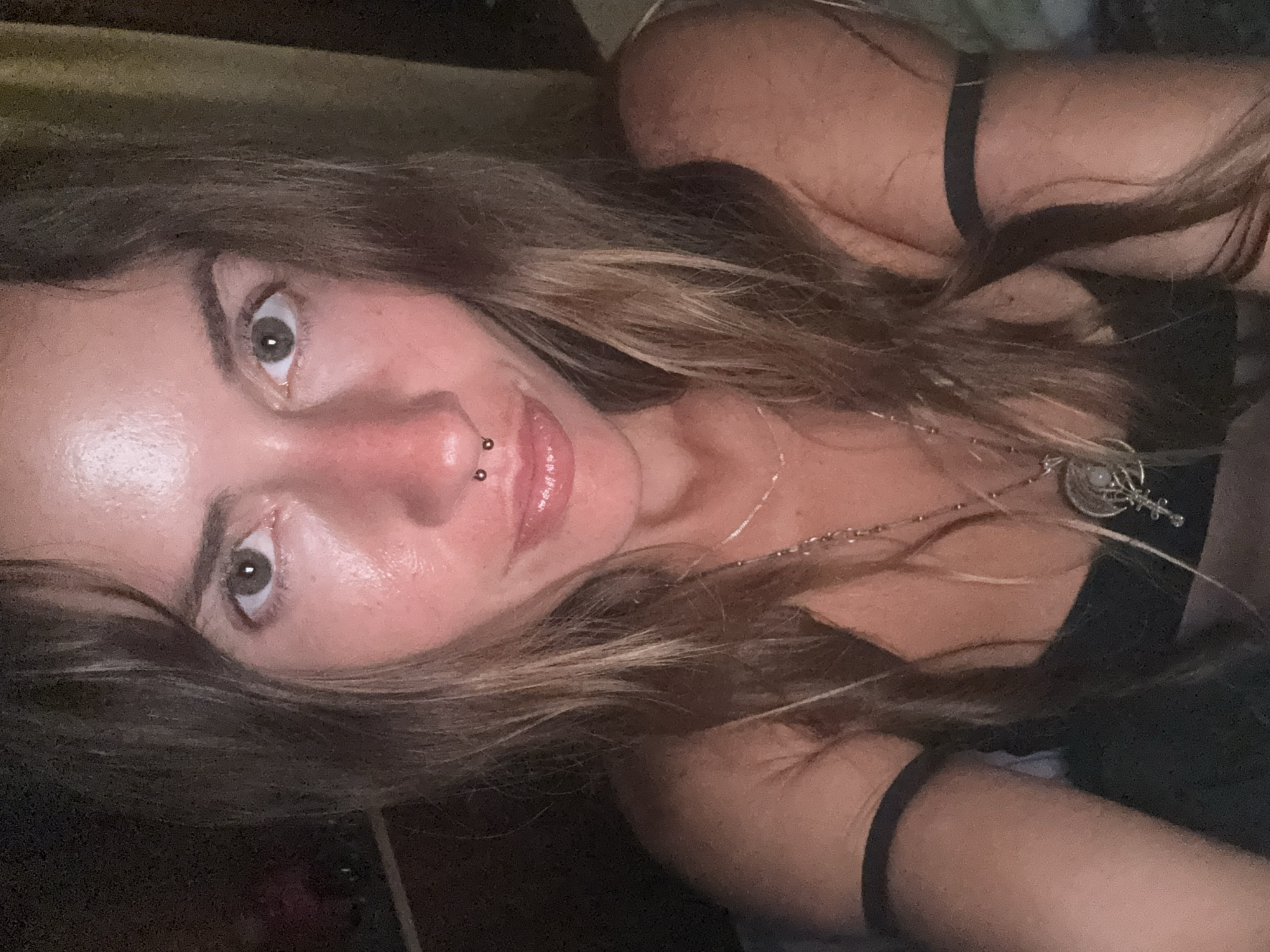 A woman with long, wavy brown hair and fair skin is shown in a close-up, frontal view. She is looking directly at the camera. She has grey-green eyes, a nose ring with two small silver balls, and her lips are slightly parted. She is wearing a dark-colored top, and a thin silver chain with a pendant is visible around her neck. Dark bands are around her upper arms. Her skin appears slightly flushed. The background is dark and indistinct, suggesting an indoor setting at night or in low light conditions. The image quality is grainy.