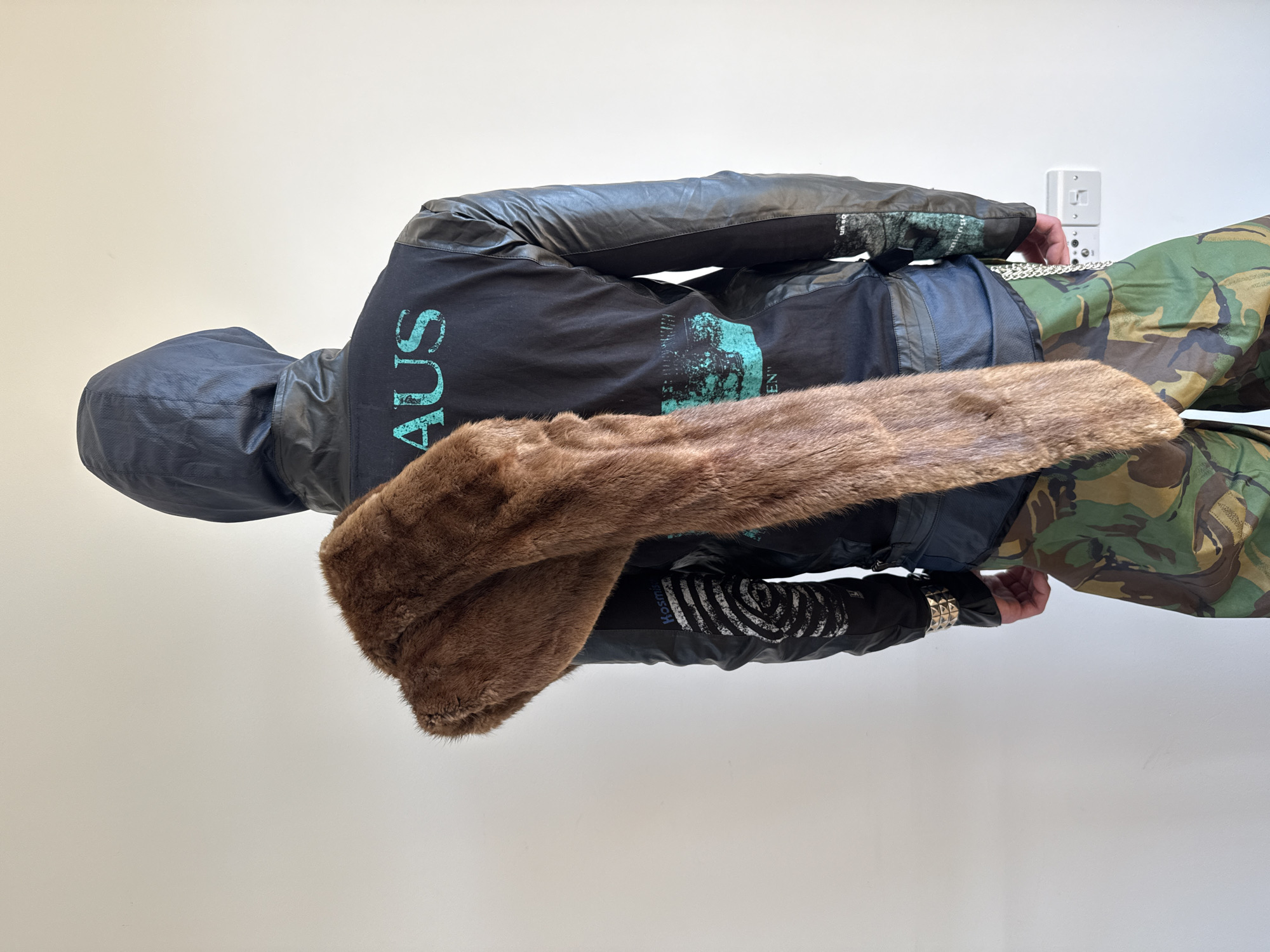 A person is captured from the back, standing against a plain, light beige wall in what appears to be an indoor setting. The individual is dressed in a distinctive, layered outfit. They wear a dark, possibly black, hooded jacket with sections that appear to be leather or a similar material, particularly on the sleeves and hood. Underneath the jacket, a black t-shirt is visible, featuring a large, vertically oriented graphic in light blue/teal on the back that reads "AUS." Below this, additional faded text and abstract designs are present, with the word "KOSMIC" partially visible on the lower left side of the back.
The person is wearing camouflage patterned pants in shades of green, brown, and black. A long, rich brown, fur-like scarf is draped around their neck, adding a textured element to the ensemble. Accessories include a silver chain hanging from the right side of their waist and a silver-studded or spiked wristband on their left wrist. A white electrical outlet with two sockets is visible on the wall to the right of the person. The individual is stationary, seemingly posing to showcase the attire. The image provides no specific cues regarding the time of day or emotions. The location context is London, United Kingdom.