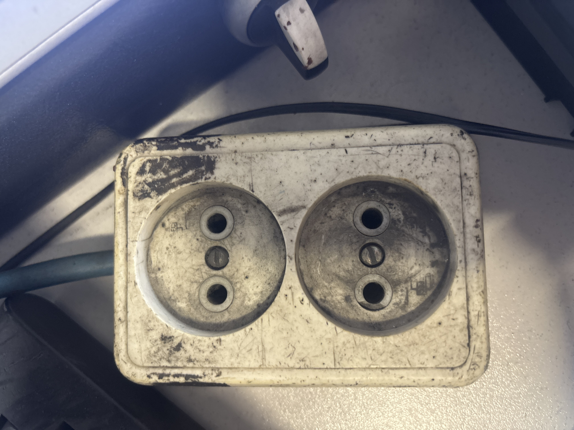 The image shows a dirty, white, double electrical socket. The socket is mounted on a textured surface and is surrounded by various dark objects, including what appears to be a black electrical device and a blue cable. The socket itself shows signs of wear and tear, with scuff marks and grime on its surface. The holes for the electrical plugs are dark and appear somewhat worn. The overall impression is one of an aged and heavily used electrical outlet. This scene is not specific to Kryvyi Rih, Ukraine, and there are no discernible elements that would indicate its location.