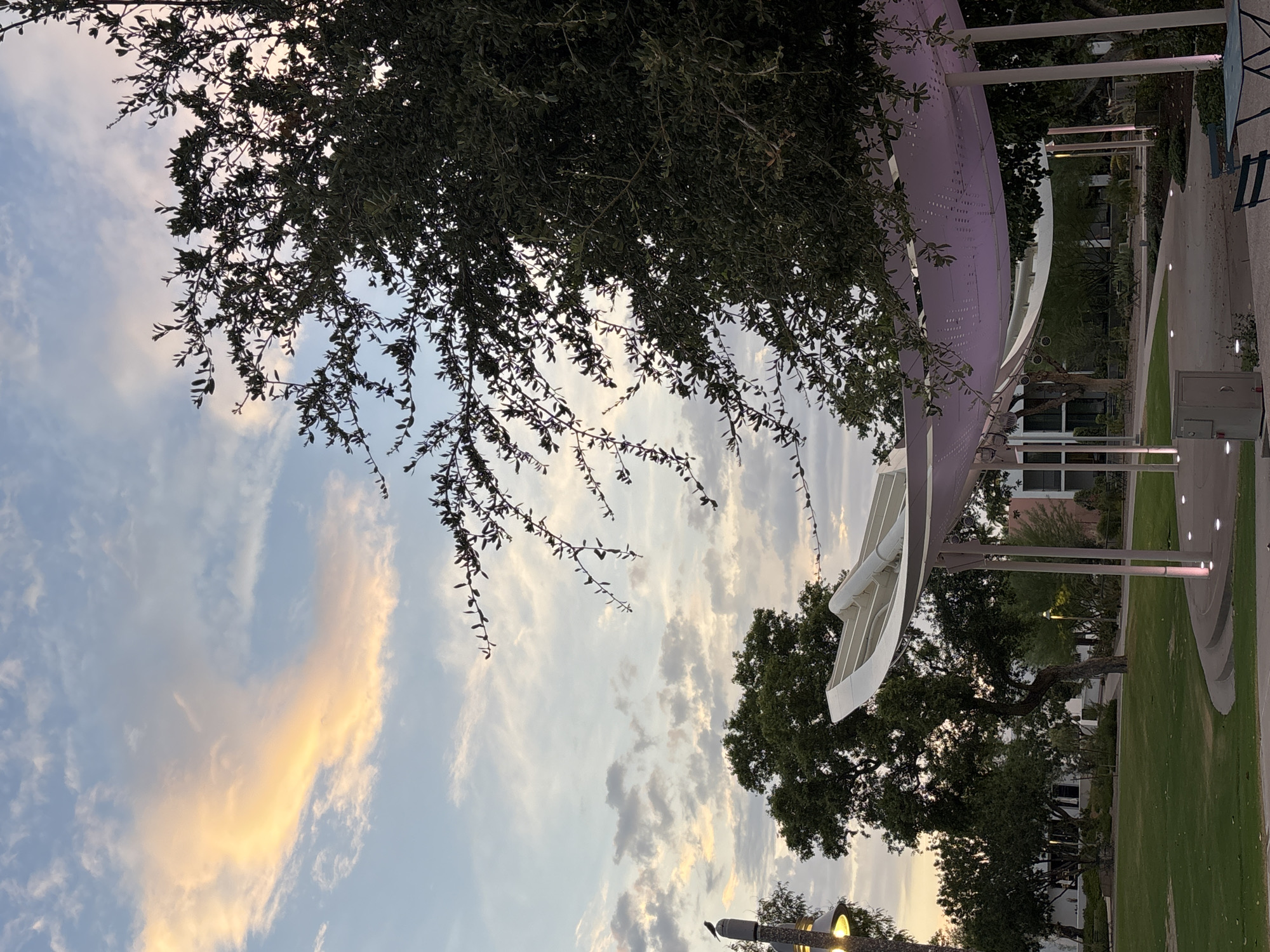 The image depicts an outdoor scene in Scottsdale, Arizona, likely during twilight. In the foreground, a curved, white-canopied structure with purple lighting is visible, supported by pillars. A small metal trash receptacle is situated to the left of this structure. Beyond the canopy, a manicured lawn slopes gently upwards, leading to various trees and buildings in the background. One prominent tree has dark green foliage and branches that extend towards the upper right of the frame. Above the trees, the sky displays a mix of clouds, with some illuminated by the soft, warm hues of the setting sun. A single bird is perched atop a lamppost on the left side of the image. The overall ambiance suggests a peaceful, urban park setting.