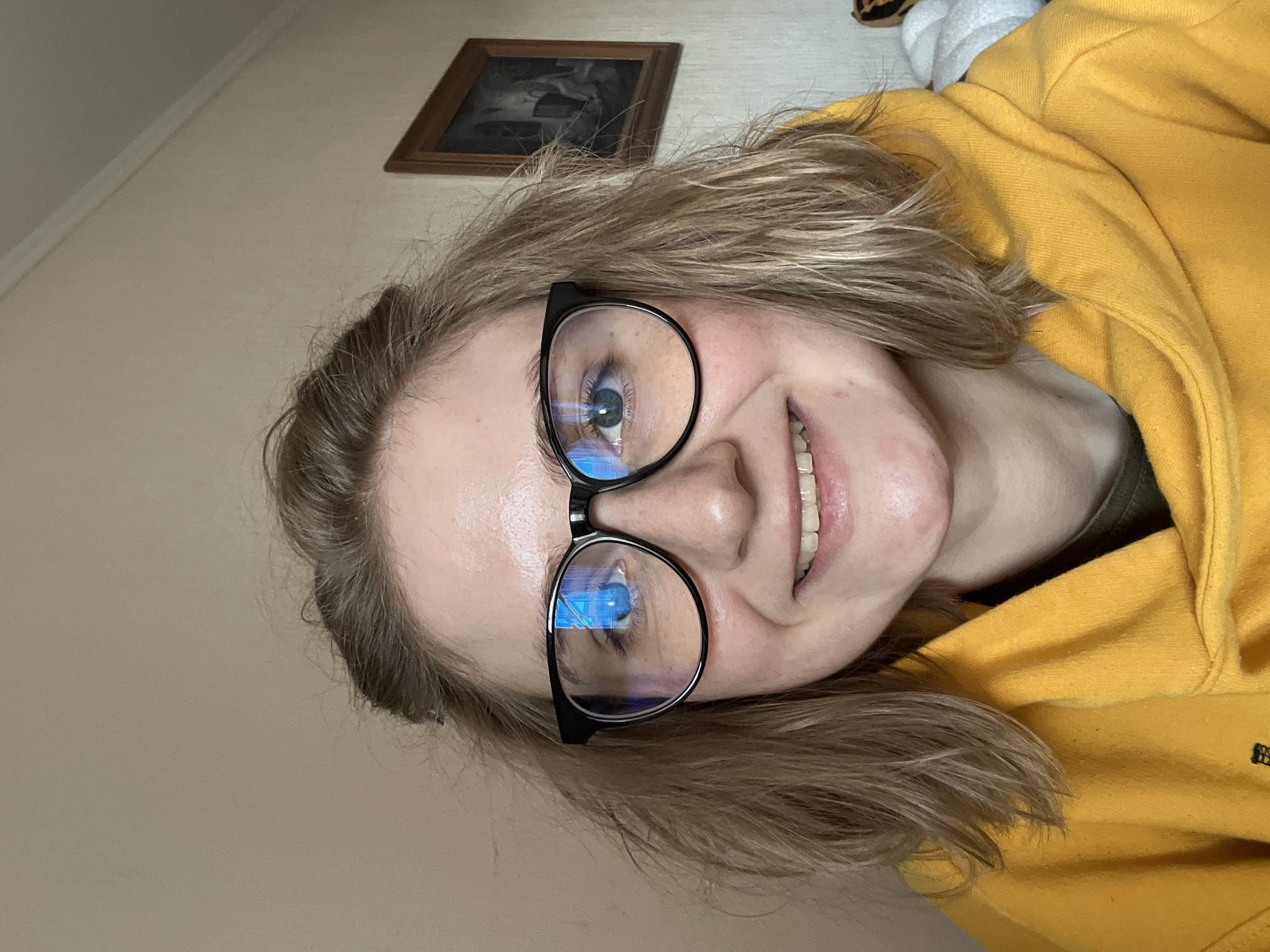 A close-up, slightly angled selfie depicts a fair-skinned person wearing black-rimmed eyeglasses and a mustard-yellow hooded sweatshirt. The person is smiling, revealing their teeth. The background includes a light beige wall with a framed picture hanging above. The reflection in the eyeglasses shows blue light. The location is not discernible as being in Novosibirsk, Russia based on the information in the image.