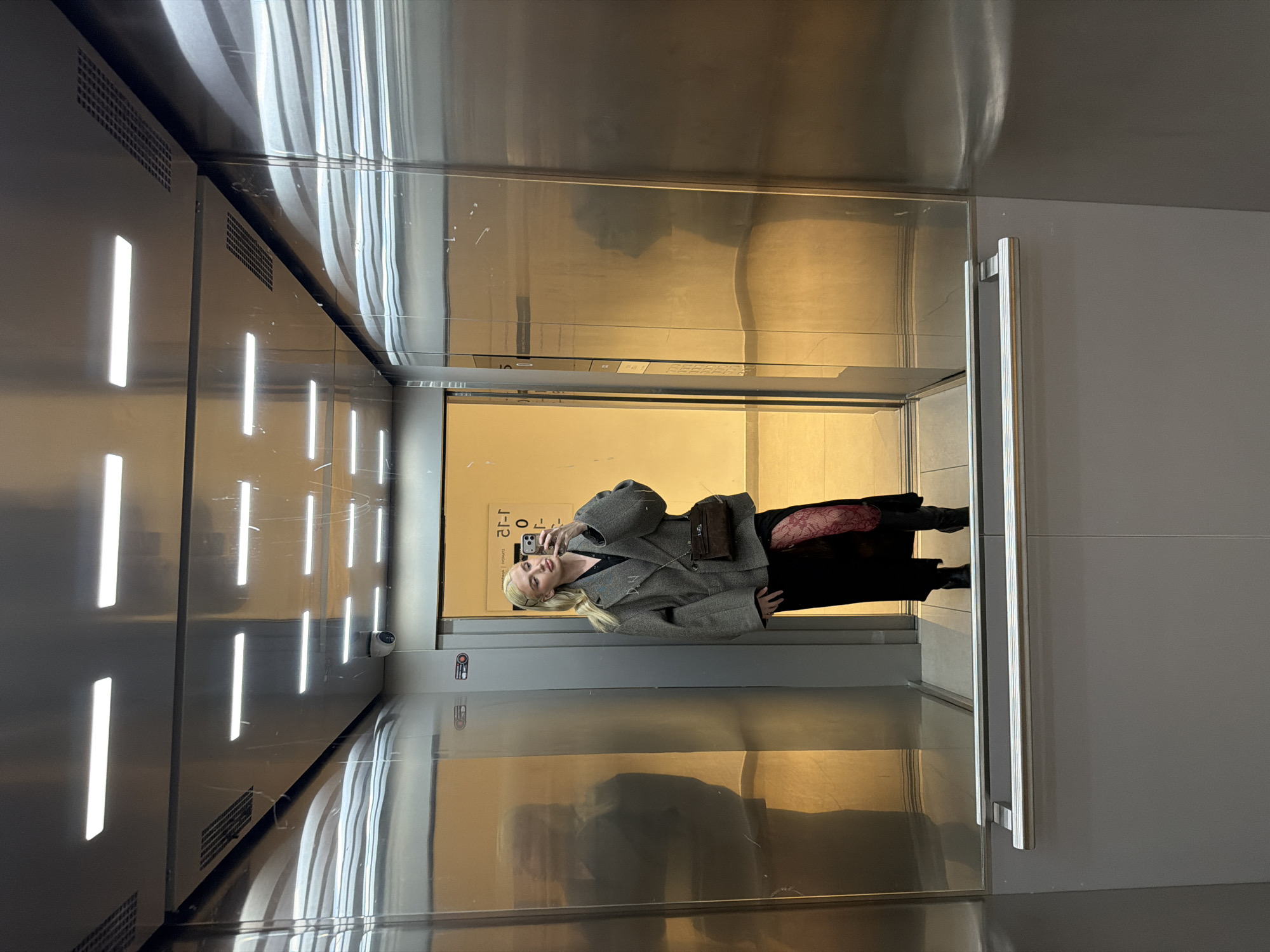 A woman stands in an elevator taking a selfie with a smartphone. The elevator has silver metal walls and a ceiling with integrated linear lighting, and a metal handle. The door is open, revealing a beige-toned space beyond. The woman wears a gray jacket, black pants, and lace stockings, and carries a shoulder bag. The reflection of the woman can be seen on the elevator walls. The city of Belgrade, Serbia, is not visible.