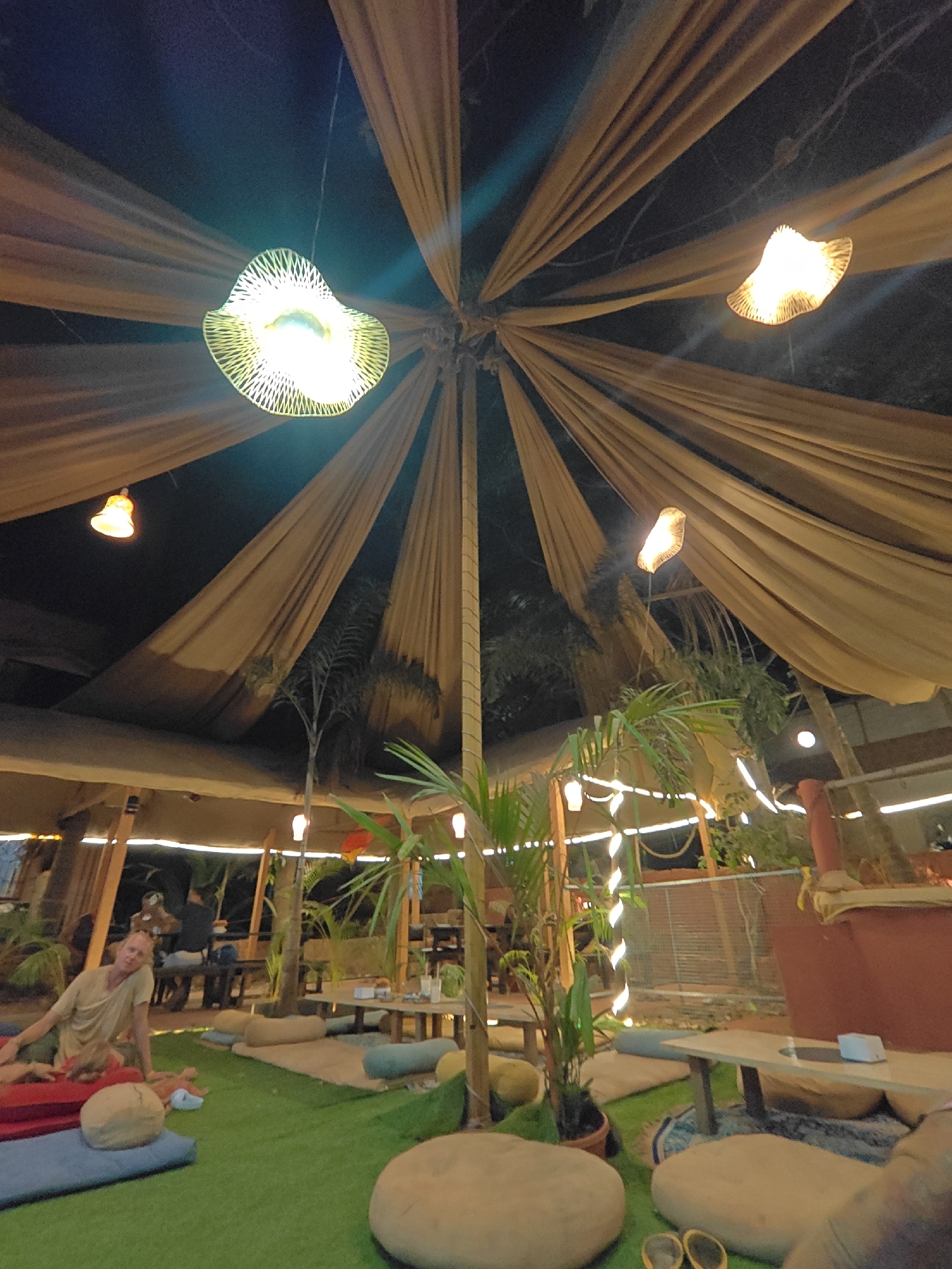 The image shows an outdoor seating area, possibly a restaurant or lounge, in the city of Unknown, Unknown. The area is covered by a tent-like structure of beige fabric, radiating from a central point. Several decorative lights hang from the fabric. Tables and cushions provide seating on a green artificial turf floor. A person is reclining on cushions in the foreground. Plants and other decorations are visible throughout the space.