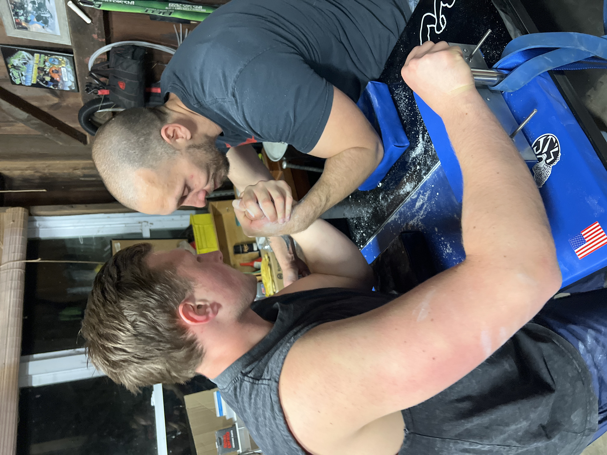 This media file captures an arm wrestling match between two men in an indoor setting, likely a garage or dedicated training area, in Orange Lake, United States of America.
The primary subjects are two men engaged in an arm wrestling contest. The man on the right has a shaved head and a beard, wearing a dark short-sleeved t-shirt. His left arm is actively competing, while his right arm holds a handle on the table. The man on the left has brown, wavy hair and wears a dark sleeveless top. His right arm is locked with his opponent's, and his left arm is braced against the table's frame. Both men show intense concentration and effort, with their gazes fixed and facial expressions indicating exertion.
The central object is a blue and black arm wrestling table, featuring padded elbow rests and a strap/handle for stability. White chalk or powder is visible on the table surface and on the men's hands and arms, used for grip. Notable details on the table include a sticker of the American flag on the blue padding and another partially visible stylized sticker.
The background suggests a functional, utilitarian space, likely a garage or workshop. Visible elements include wooden shelves, framed pictures on a wall, a long green object (possibly a tool or sports equipment), and various stored items including cardboard boxes. The lighting appears to be artificial, typical of an indoor, enclosed space, making the time of day undeterminable. The overall atmosphere is one of focused athletic competition.