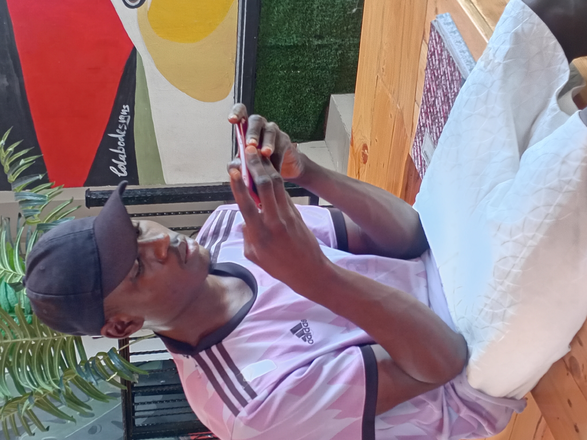 A young Black male with dark skin and a short haircut, wearing a black baseball cap, a pink Adidas t-shirt with black stripes on the sleeves, and white shorts, is seated on a wooden bench and looking down at a red smartphone he is holding with both hands. He is positioned in the foreground, facing slightly to his right. Behind him, on the wall, is a colorful mural with abstract shapes in red, black, green, beige, and yellow, and the text "Colabodesigns" is visible in white cursive lettering. To the left of the male, a large green plant with striped leaves is visible. The setting appears to be an outdoor seating area, possibly a cafe or restaurant, with a wooden table in front of the seated male. The overall lighting is bright and natural.