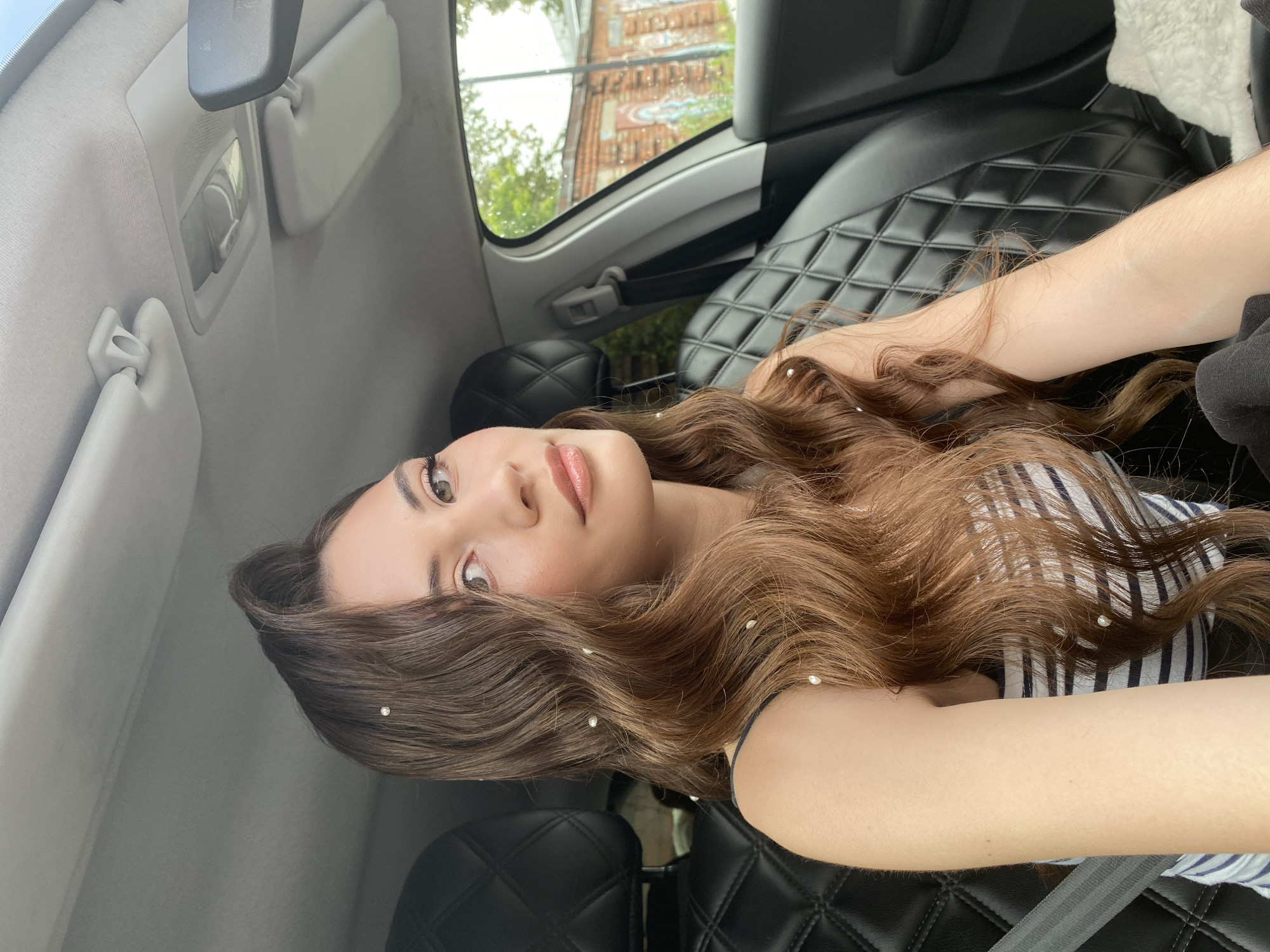 A young woman with long, wavy brown hair adorned with small pearls sits in the driver's seat of a car. She is wearing a striped tank top and has light makeup. Her hair is styled with volume and loose curls, with several pearls embedded throughout. The car's interior is visible, with black leather seats featuring a diamond pattern and a grey ceiling. A seatbelt is fastened across her shoulder. Through the car window, an outdoor scene can be seen, including a brick building with graffiti on it and some greenery, suggesting an urban or suburban environment. The lighting indicates it is daytime. Based on the style of the car and the surrounding architecture, the scene is likely in Votkinsk, Russia.