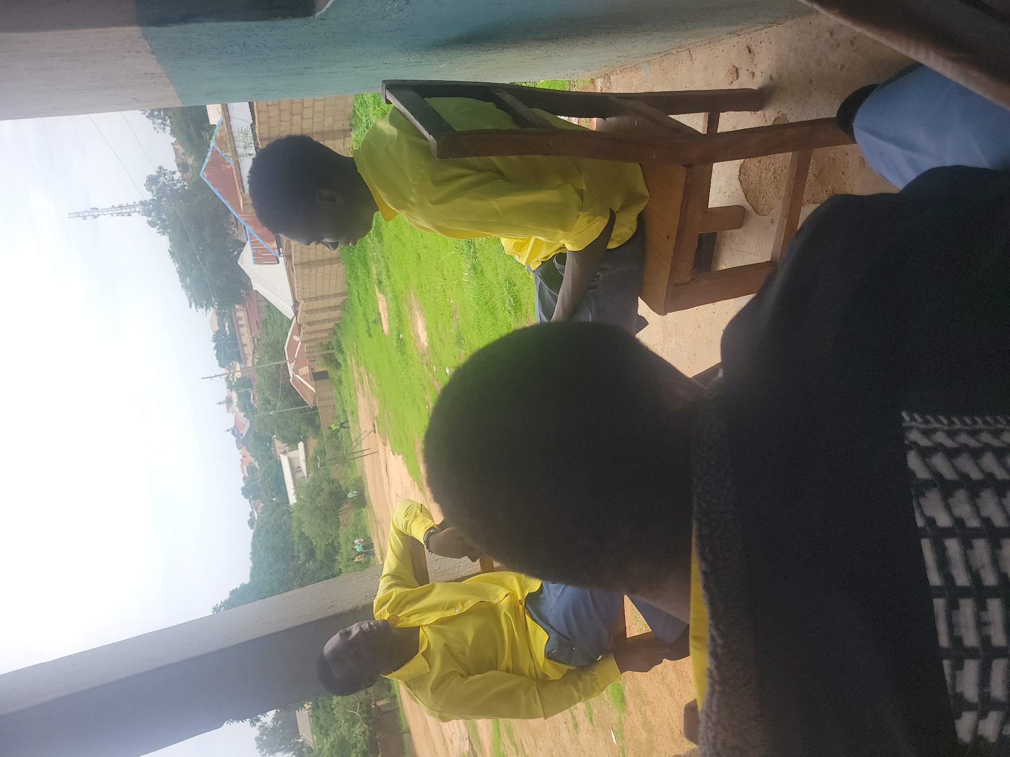 The image shows several people in an outdoor setting in Jos, Nigeria. Two young people in yellow collared shirts are seated on chairs. One young man is turned away, partially visible in the foreground wearing a black garment. There's a grassy area visible behind the young people and buildings, including residential structures, with a communications tower in the distance. The area appears to be an educational setting with an outdoor study location or a community gathering space.