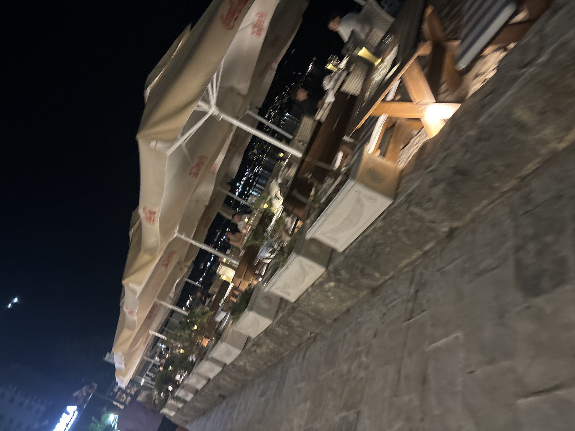 The scene is an outdoor restaurant in Budva, Montenegro, at night. The restaurant stretches along a stone walkway, lined with numerous tables under Coca-Cola branded umbrellas. Patrons are seated at some tables, though details about their activities are limited due to the image's blur and angle. White planters, some containing greenery, are situated between the tables and the walkway's edge. The background features city lights reflecting off the water. The photograph has a tilted perspective and is blurry, making details difficult to discern.