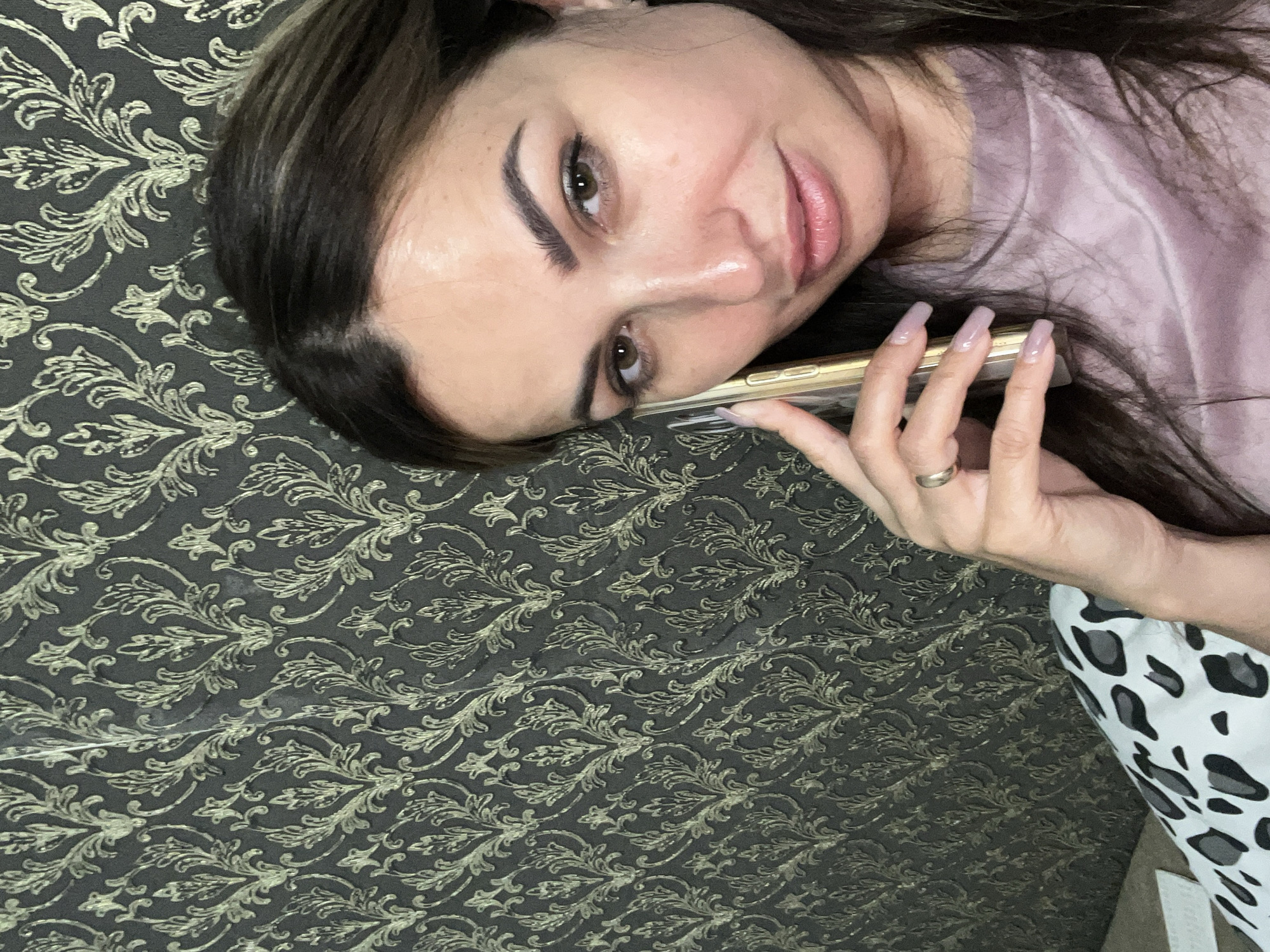 A medium shot shows a woman with dark hair and light brown eyes holding a gold smartphone to her left ear. She is looking towards the camera with her head tilted slightly to the right. The woman is wearing a light purple shirt and a ring on her left ring finger. The background is a dark gray wallpaper with a gold ornate damask pattern. To the lower left of the image, a portion of a pillow with a black and white leopard print is visible. The lighting appears to be artificial and is coming from the front and slightly to the left of the woman.