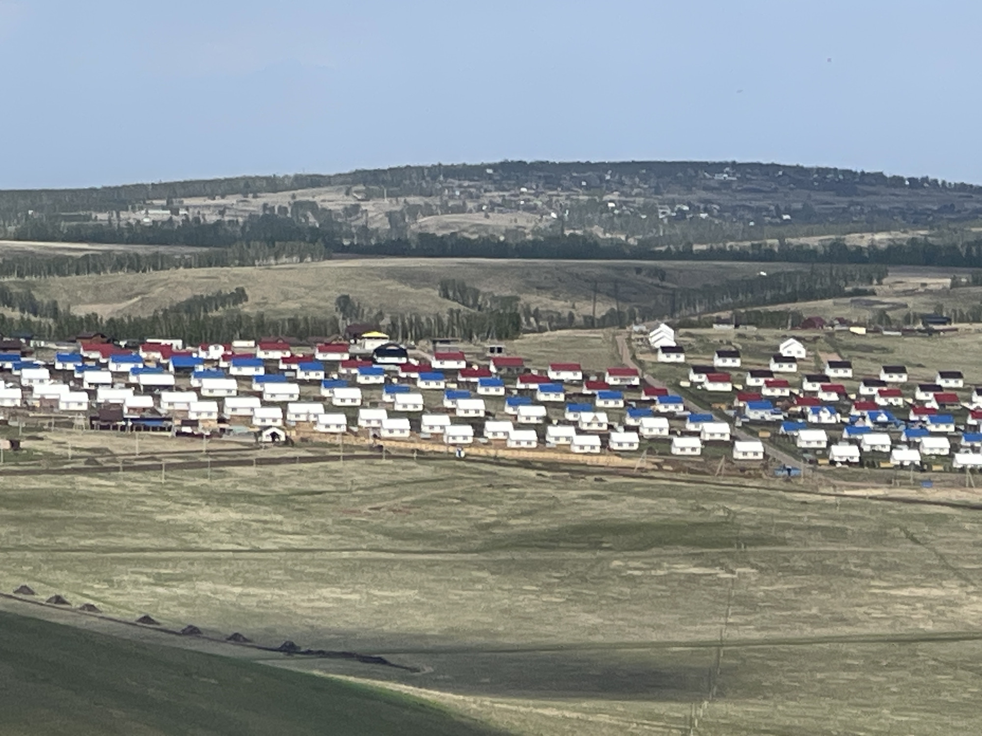 A high-angle, long shot depicts a housing development near Солонцы, Russia. The development is situated in a grassy, rural area, with a line of trees and a hill in the background. The houses are small, white, and seemingly identical, with roofs painted in alternating colors of red and blue. There are multiple rows of houses spread throughout the area. A single building has a yellow roof, which is towards the center of the composition. The foreground is a field with patches of greenery and evidence of soil disturbance.