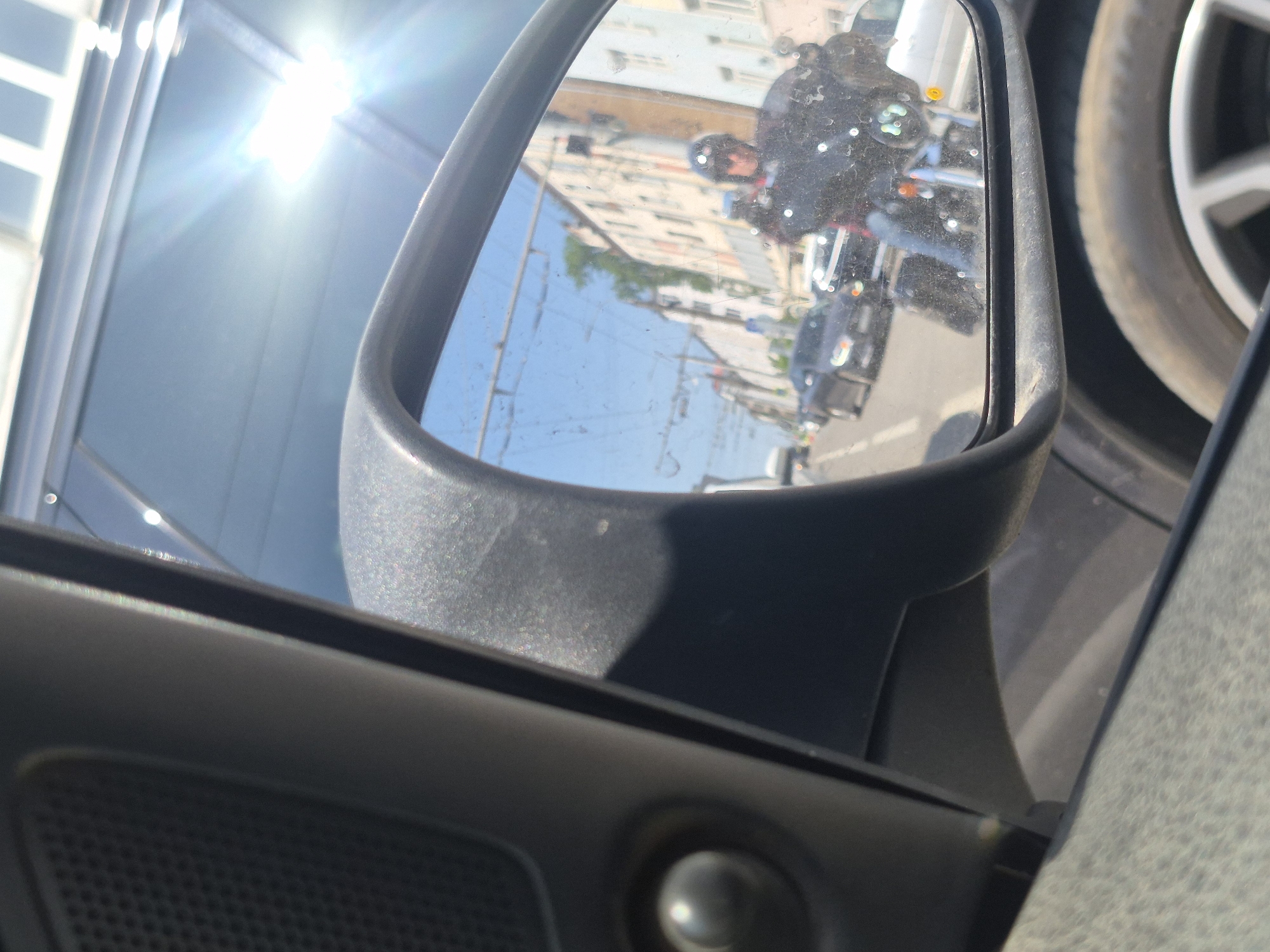 A side mirror reflects a street with buildings and a motorcycle, with a rider wearing a helmet. The motorcycle is directly behind the car with the mirror. The mirror is attached to a dark-colored car. A wheel and tire of the car is visible in the upper right of the frame.