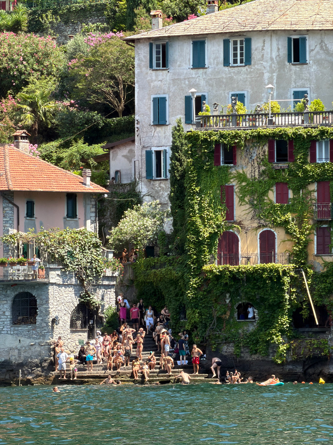 The image depicts a lively scene at a lakeside public area in Nesso, Italy. In the foreground, the rippling green water of Lake Como is visible, with several individuals swimming and relaxing. A person on an orange inflatable float is positioned in the mid-ground of the water.
The focal point of the image is a stone staircase leading from the water up to buildings adorned with lush green ivy. A large crowd of people, dressed in swimwear, are seated and standing on these steps, socializing and enjoying the sunny weather. Some are interacting with each other, while others are looking out at the lake.
Behind the steps, two buildings are prominently featured. The building on the left has a reddish-brown tiled roof and light-colored walls, with arched windows. The larger building on the right is a multi-story structure covered in dense green ivy. It has multiple windows with dark shutters and a balcony overlooking the lake, where a few people are visible. The architectural style suggests historic European lakeside residences. The background consists of a steep, verdant hillside covered in trees and flowering bushes, characteristic of the Italian lake region. The overall atmosphere is one of a popular summer gathering spot.