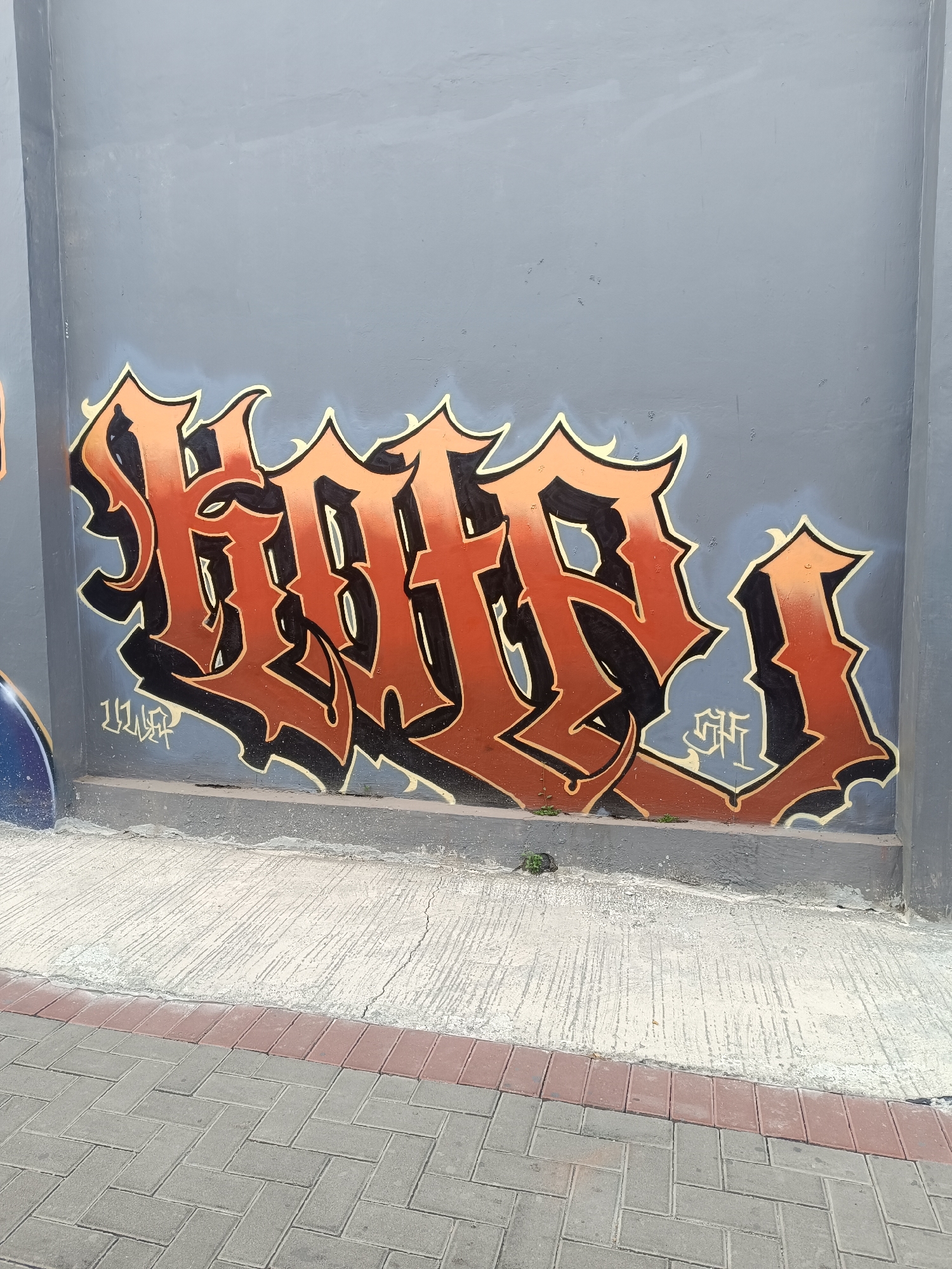 In Yogyakarta, Indonesia, a large graffiti piece featuring bold, orange and black stylized lettering adorns a grey concrete wall. The graffiti spans across the wall, its sharp angles and dramatic shading giving it a dynamic, almost three-dimensional appearance. Below the main artwork, smaller tags are visible, one in white with black outlines and another in light blue with black outlines. The wall itself shows signs of wear, with chipped paint and some minor damage near the base. In front of the wall is a paved walkway, transitioning from cracked concrete to interlocking grey and red bricks. Small green plants are growing at the base of the wall, pushing through cracks in the concrete. The lighting suggests it is daytime.
