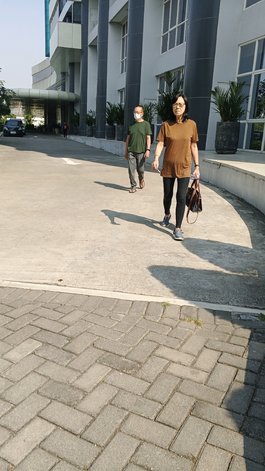 Two individuals, an older man and a woman, are walking on a paved outdoor area in front of a modern building. The man, wearing a green t-shirt, gray pants, and sandals, has a white mask covering his nose and mouth. The woman, in a brown t-shirt and black leggings, is carrying a dark-colored bag and a bottle of water. The building behind them features large pillars and windows, with potted plants visible along its facade. The ground is a combination of concrete and brick pavers. The lighting suggests it is daytime, with long shadows cast by the individuals. A black car is parked to the left of the frame, near the entrance of the building. The overall scene appears to be a public area, possibly a walkway or courtyard of a commercial or institutional building. There is no direct indication that this location is in Semarang, Indonesia; however, the architectural style and vegetation could be consistent with urban environments in Southeast Asia.