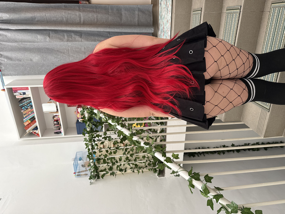 The image displays a person from the back, captured in an indoor setting, apparently standing on or next to a staircase. The image itself is rotated 90 degrees clockwise, so the description will assume an upright orientation for clarity.
The central subject is a person with very long, thick, and vibrant bright red hair that cascades down their back. They are wearing a black pleated mini-skirt, which features several silver or white eyelets or grommets along the pleats. Beneath the skirt, they are wearing black fishnet stockings, which are visible on their thighs and lower legs. Over the fishnet stockings, they have on black knee-high socks with two horizontal white stripes near the top cuff. Their bare back and upper arms are visible above the skirt.
The setting appears to be a residential or multi-level indoor space. To the right of the person (if the image were upright), there is a staircase with light-colored, possibly white, railings. Green artificial ivy garlands are wrapped around these railings, adding a decorative element. The stair treads have a patterned or tiled surface.
In the background, to the left of the person (again, if upright), there is a white bookshelf filled with numerous books, some stacked horizontally and others standing vertically. The book titles are not clearly legible. Next to this larger bookshelf, a smaller white shelving unit or cabinet is visible, holding various items. Further to the left and lower in the frame, a small, light-colored birdcage containing at least one small bird is visible on a surface. Above the bookshelf, a light grey or white curtain or blind partially covers a window or wall. The overall lighting is bright and even, suggesting it is daytime. No specific activity beyond standing is discernible. No clear text is visible in the image apart from the unreadable book titles on the shelves.