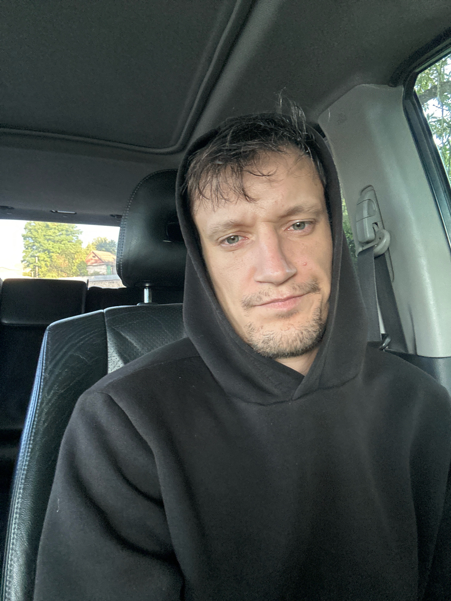 A man, appearing to be in his late 20s or early 30s, is captured in a selfie taken from inside a car. He is wearing a black hoodie with the hood up, partially obscuring his face. The man has fair skin, light-colored eyes, and short, messy brown hair. He has a light stubble on his chin and jawline, and a small mole is visible on his right cheek. His expression is neutral, with a slight upward tilt of his head. The interior of the car is visible, including black leather seats and the car's roof lining. Outside the car window, a rural landscape can be seen, featuring green trees and a building with a red roof, consistent with the environment of Makiv, Ukraine. The lighting suggests it is daytime, possibly late afternoon, given the shadows and the quality of light. The overall scene is casual and personal, indicating the man is likely the driver or a passenger in the vehicle.
