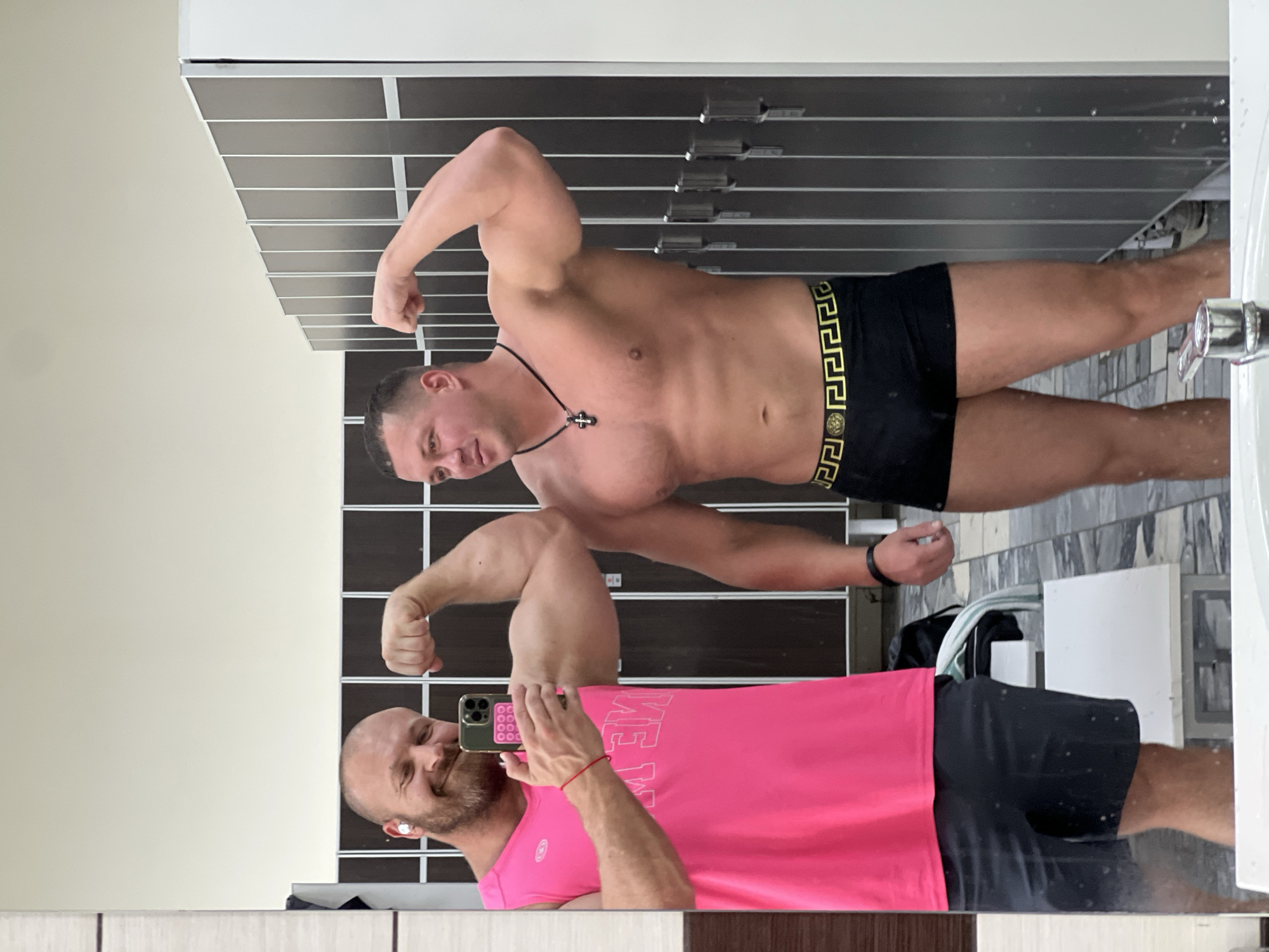 In a locker room setting, two men are captured in a mirrored reflection. The man in the foreground, seen from the waist up, is bald with a beard and is wearing a bright pink tank top. He holds a smartphone in his left hand, taking the photo, and has a small red string bracelet on his left wrist. He appears to be smiling.
Behind him, reflected in the mirror, is another man. He is flexing his bicep and is wearing black athletic shorts with a gold patterned waistband, resembling Versace. He is also wearing a necklace with a cross pendant. The background of the reflection shows dark-colored lockers. The overall lighting suggests an indoor environment, possibly a gym or sports facility. The presence of lockers and the tiled floor in the reflected area indicates a locker room or changing area. The image appears to be taken during daylight hours, given the ambient light.