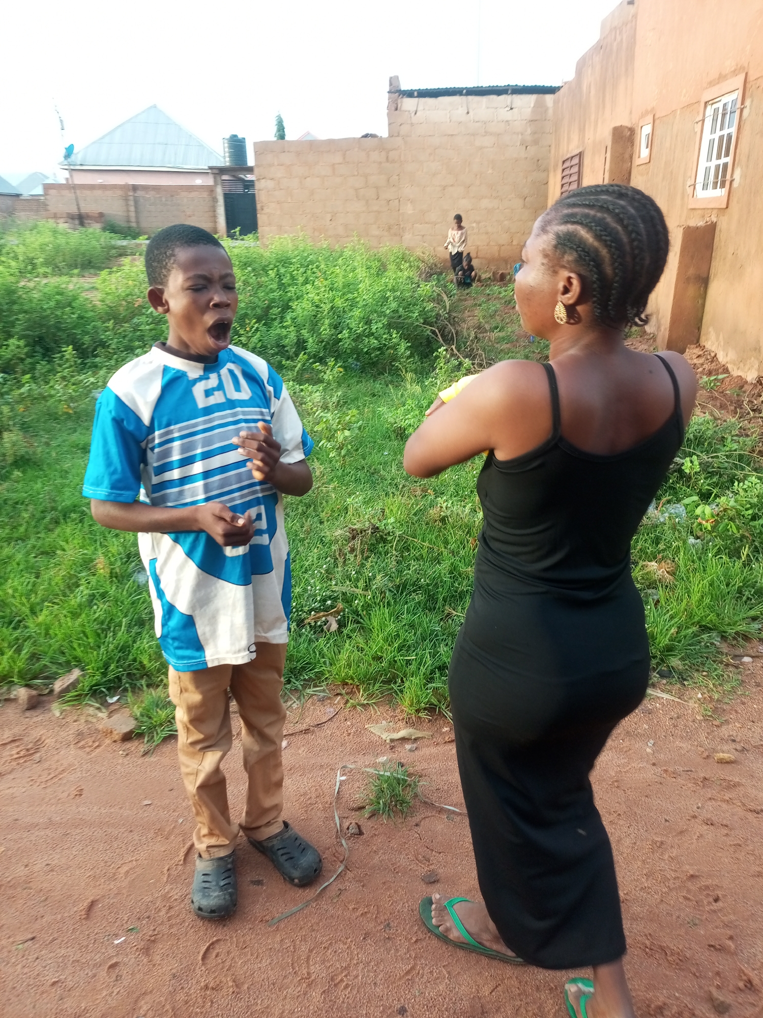 In Tempure, Nigeria, a young boy in a blue and white striped t-shirt and brown pants, wearing black clogs, is captured mid-yawn while standing on a dirt path. Facing him, a woman in a long black dress, adorned with hoop earrings, stands with her back to the camera. Green vegetation and brick buildings, typical of the urban landscape, are visible in the background. The scene appears to be outdoors, possibly in a residential area, with an overcast sky suggesting a potentially humid atmosphere.