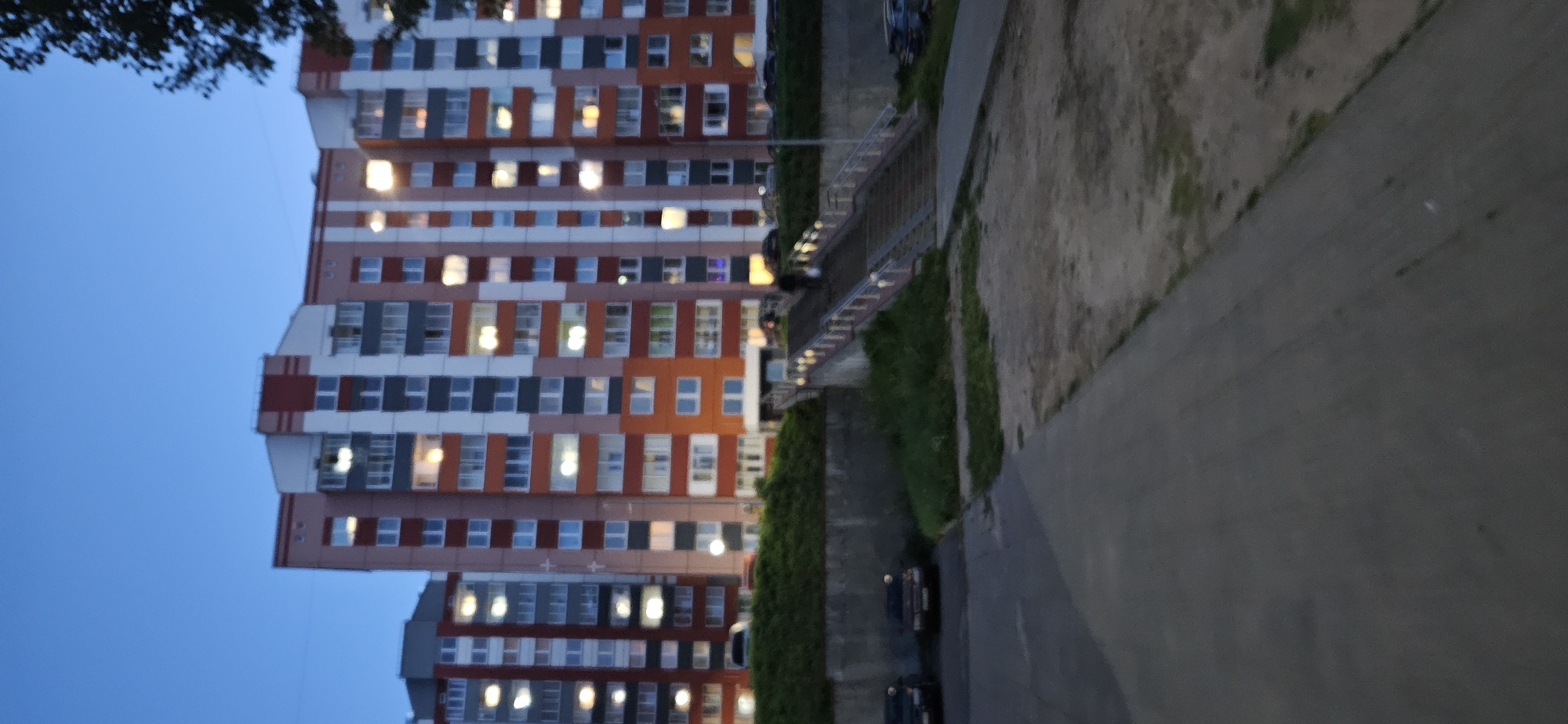 The image depicts a twilight scene in a residential area, likely in Vitsebsk, Belarus, given the architectural style of the apartment buildings which is common in post-Soviet countries. In the background, several multi-story apartment buildings with colorful facades of orange, pink, white, and grey are visible. Many windows are illuminated, suggesting occupancy. In the mid-ground, a concrete retaining wall with a grassy slope on top separates the street level from the buildings. A wide concrete staircase with metal railings and lights on either side leads up towards the buildings. One person, appearing as a dark silhouette, is ascending the stairs carrying a bag. In the foreground, a paved walkway or road is visible, with several parked cars on the left side of the image. The sky is a deep blue, indicating it is either dusk or dawn. The overall impression is of a typical urban residential environment.