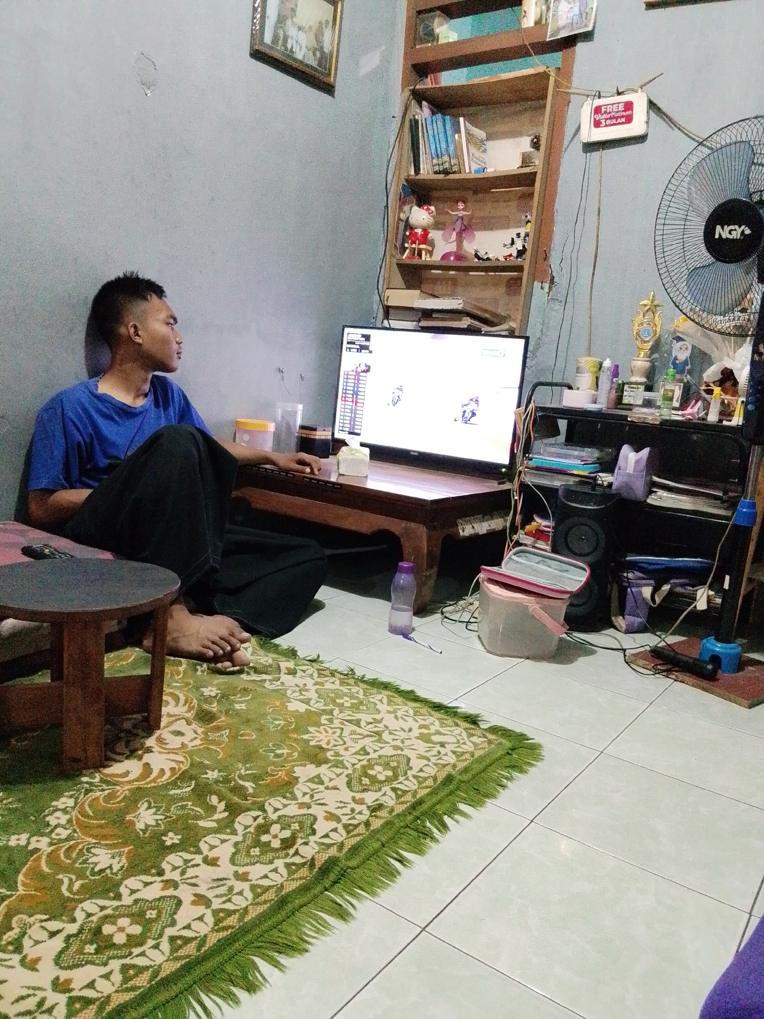 A young man sits on the floor, facing a television screen, in a room that appears to be in Pasuruan, Indonesia. The room is sparsely furnished, with a wooden shelf unit against the wall holding books and decorative items, and a small wooden table in front of the television. The man is wearing a blue t-shirt and dark pants, and he is wrapped in a dark fabric. A green and cream patterned rug covers part of the tiled floor. A standing fan is visible in the background, along with various electronic devices and cables. A framed picture hangs on the wall above the shelf. The overall impression is of a modest living space.