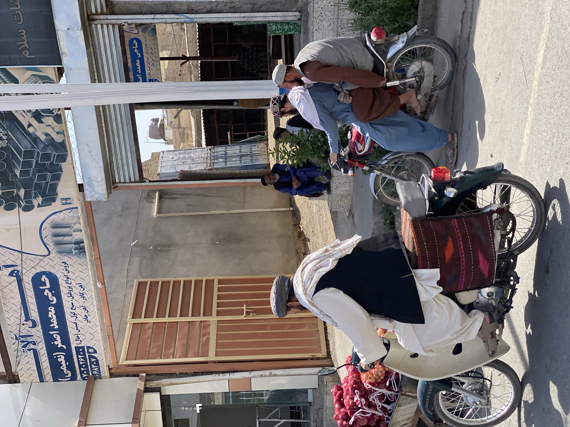 In an outdoor market scene in Kandahar, Afghanistan, two men ride on a motorcycle, with one operating the vehicle and the other seated behind him. The motorcycle is a red Honda model. The man driving wears a light blue shalwar kameez and a white turban, while the passenger is dressed in a brown vest over a grey shalwar kameez and wears a grey cap. They are positioned on a paved street, with buildings and shops lining the background. To the left of the frame, another motorcycle is parked, carrying a large load of red onions in a wooden cart. This motorcycle is cream-colored with green accents. The man on this motorcycle is visible from the chest down, wearing a black vest over a white shalwar kameez and a checkered turban. He is facing away from the camera. In the background, several other individuals are visible, some sitting on steps and others walking. The architecture suggests a typical Afghan urban setting, with storefronts and metallic roll-up doors. The overall atmosphere is that of a bustling marketplace on a sunny day.