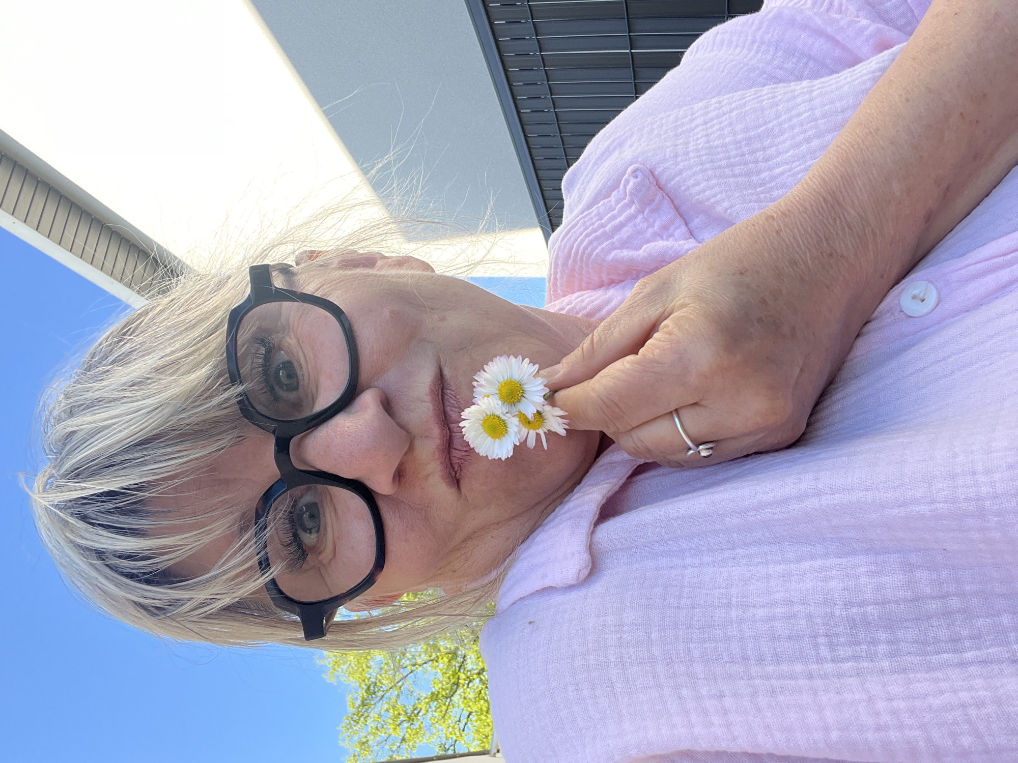 A medium shot presents an older woman with short, gray hair and wearing large, round, black-rimmed eyeglasses. She's positioned outdoors with a pink, button-up shirt. She holds three small white daisies with yellow centers near her face. The backdrop includes a clear blue sky, green tree foliage, and the architecture of a building with both a white exterior and dark gray accents. The photograph was taken in Chambœuf, France.