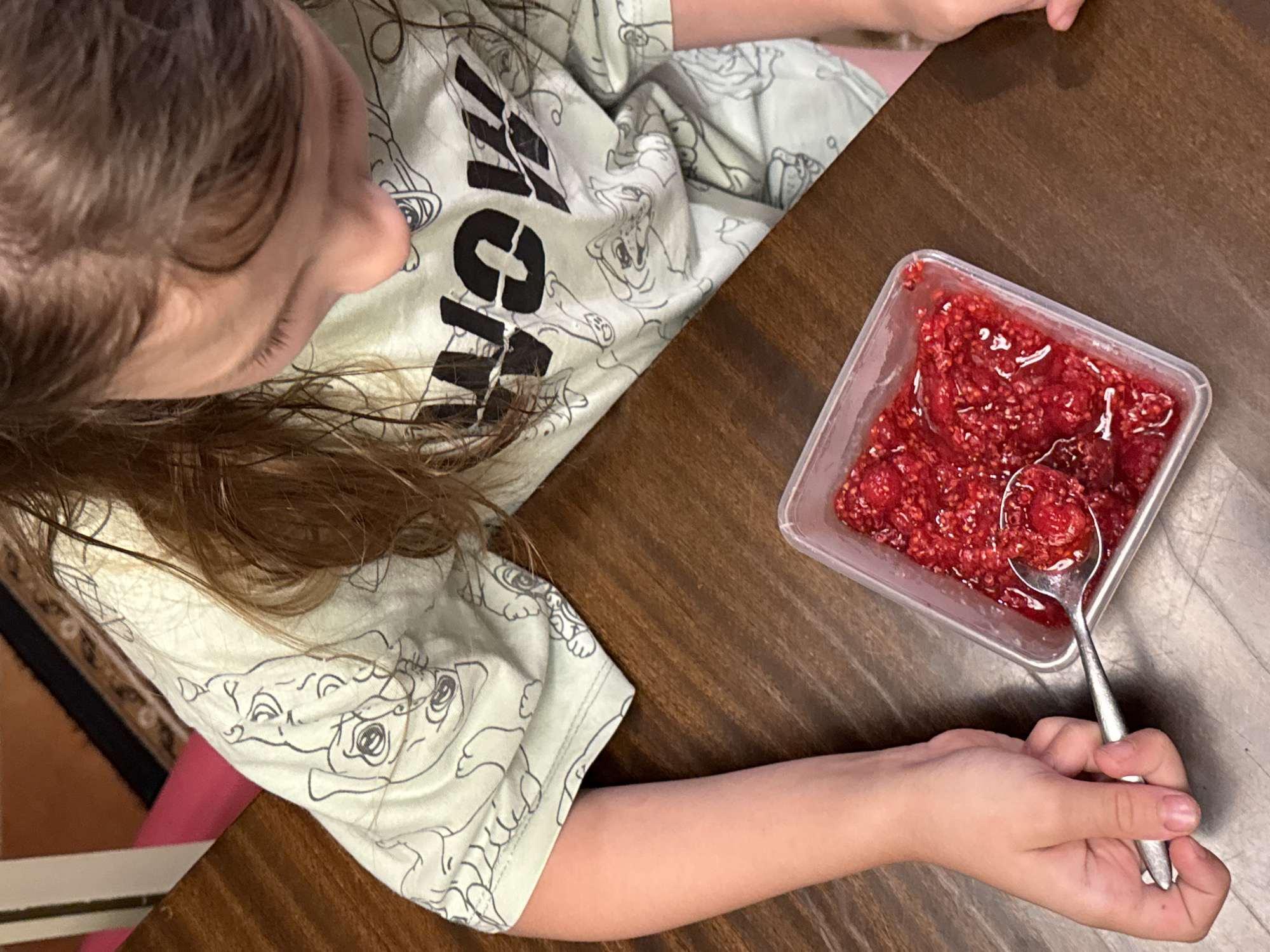 In this overhead shot, a young person with light brown hair is seated at a dark wood table. The child is wearing a light green t-shirt adorned with a repeating pattern of cartoon characters, some of which are black outlines of faces. The child's left arm rests on the table, while their right hand holds a spoon filled with a red, chunky substance, likely jam or fruit preserves. The spoon is positioned above a clear plastic container also filled with the same red substance. The scene appears to be indoors, with the lighting suggesting it could be daytime. The overall mood is casual and domestic. There is no visible text in the image. The location context is Zassovskaya, Russia.