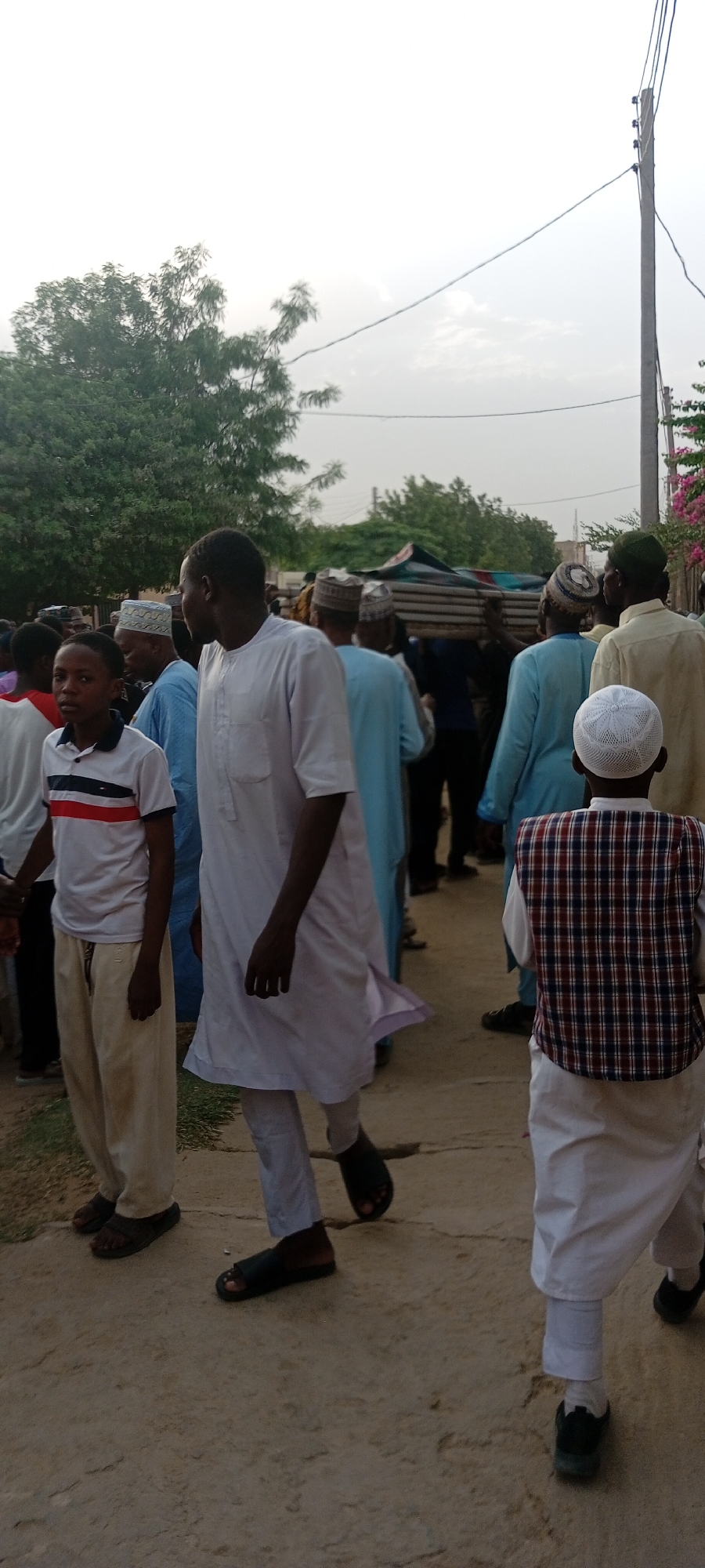 The image, taken in Sabon Gari West, Nigeria, shows a group of people gathered outdoors, seemingly walking together along a street. The group includes men and boys of various ages. Most of the men are wearing traditional Muslim attire, including robes and caps. One man is wearing a white robe. Others are wearing blue robes. A boy to the left is wearing a white t-shirt with red and black stripes, and tan pants. Another boy to the right has a checkered vest over a white outfit, along with a cap. The ground is a dusty, paved surface. In the background, there are trees, an electric pole, and possibly buildings. Some members of the group appear to be carrying an object.