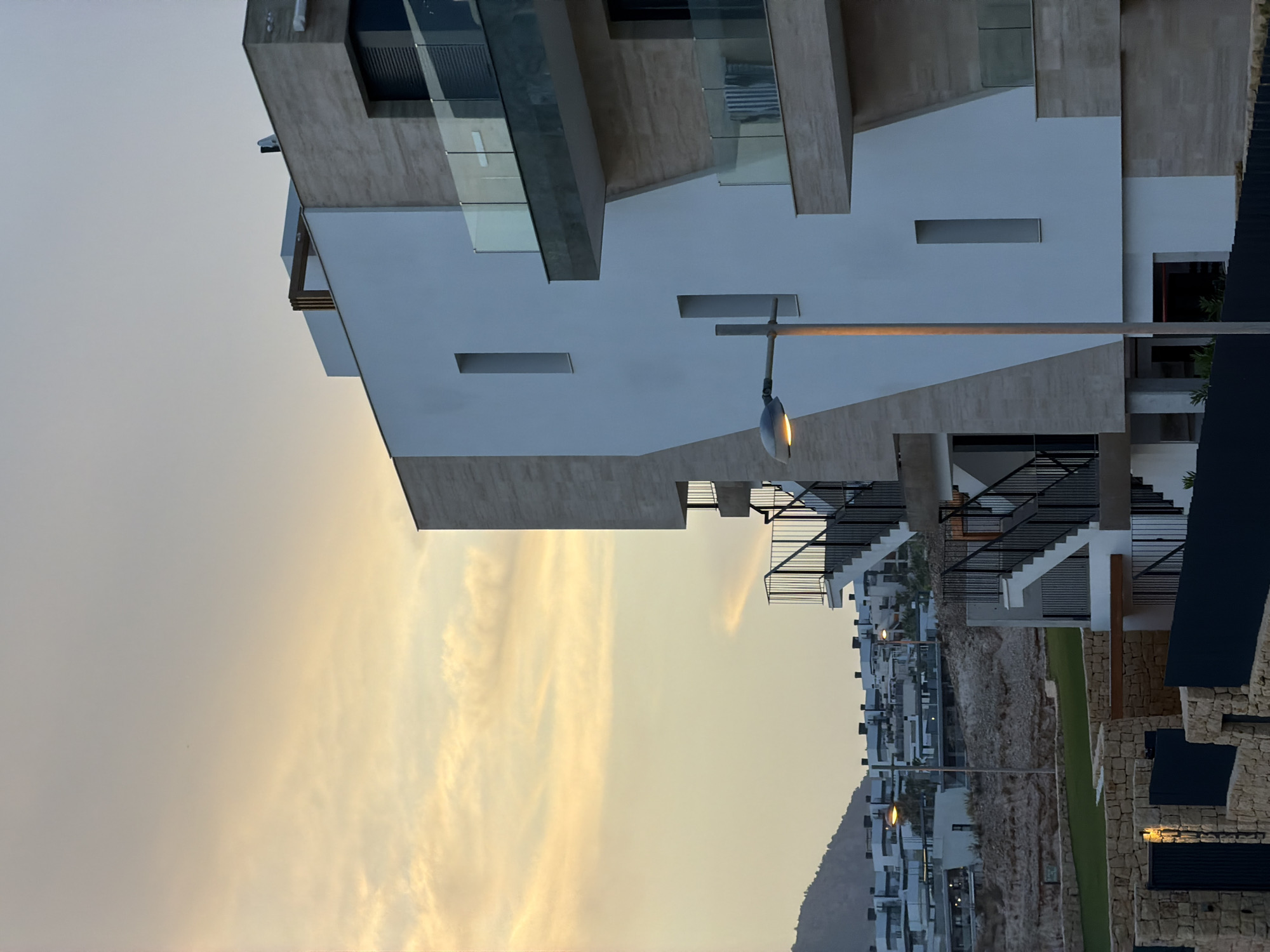 This is a daytime, outdoor, medium shot photograph taken in Finestrat, Spain, showing a modern apartment building under a sky with scattered clouds tinged with yellow and orange hues, suggesting either sunrise or sunset. The building features a facade of white panels and light-colored stone, with balconies adorned with glass railings. To the left, a hill is visible with numerous white buildings constructed on its slope. In the foreground, a low stone wall with dark metal fencing and a lit lamppost are present. The image captures an urban architectural scene with a natural backdrop.