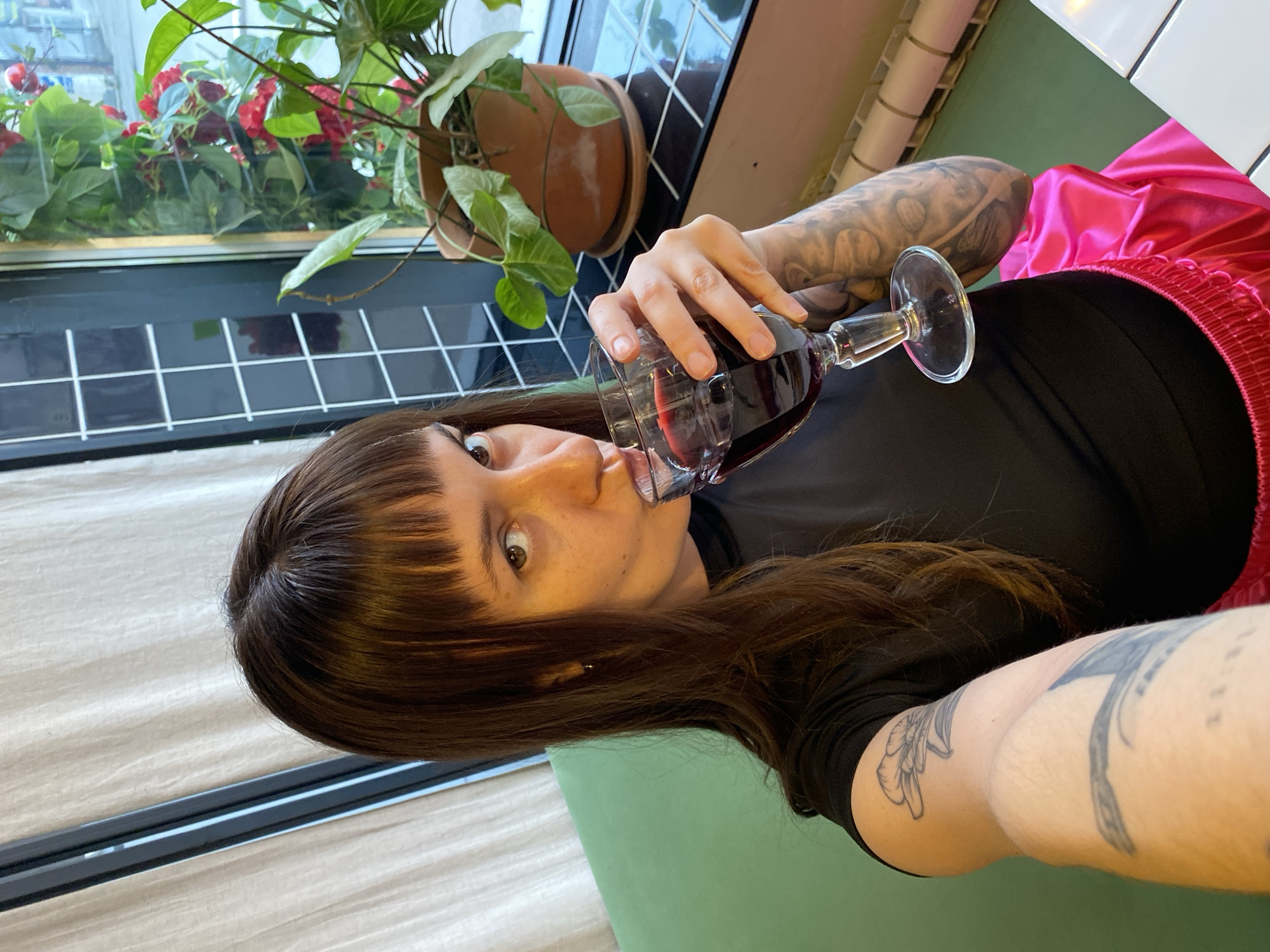 A medium shot depicts a woman with brown hair and a fringe, drinking from a wine glass filled with red wine. She is wearing a black top and pink shorts with a red trim. The woman's left arm, visible in the foreground, is adorned with tattoos. In the background, there's a window with a view of greenery and flowers, as well as black and white checkered tiles. The woman sits on a green surface. Location is Votkinsk, Russia.
