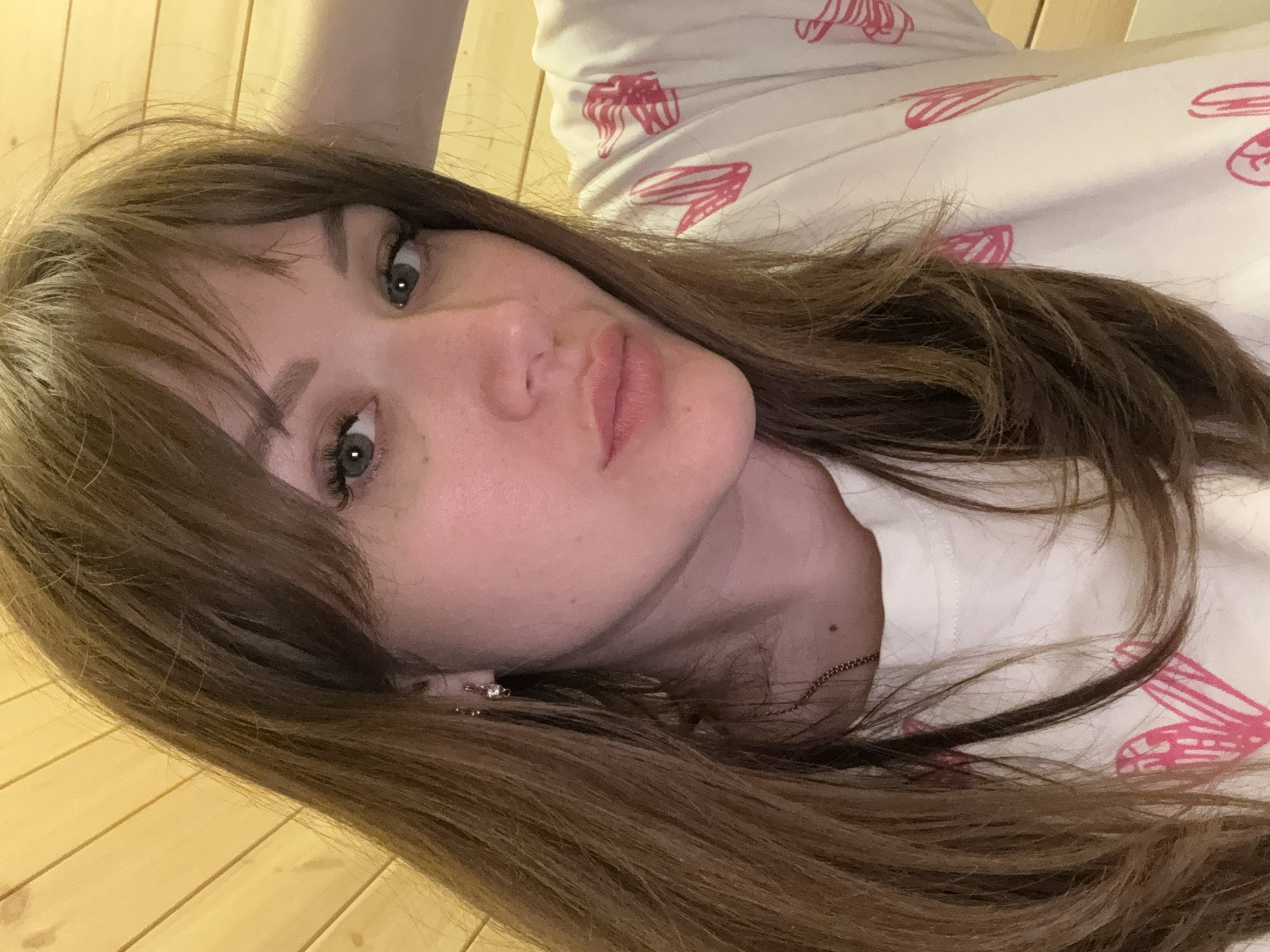 A close-up portrait of a woman with light skin, brown hair, and green eyes. She is lying down, and the camera angle suggests she is looking up. Her hair is styled with bangs and appears to be layered. She has subtle makeup on. She is wearing a white t-shirt with pink prints on it. The background includes wooden planks and part of a patterned fabric. There is no indication of the city of Taman', Russia.