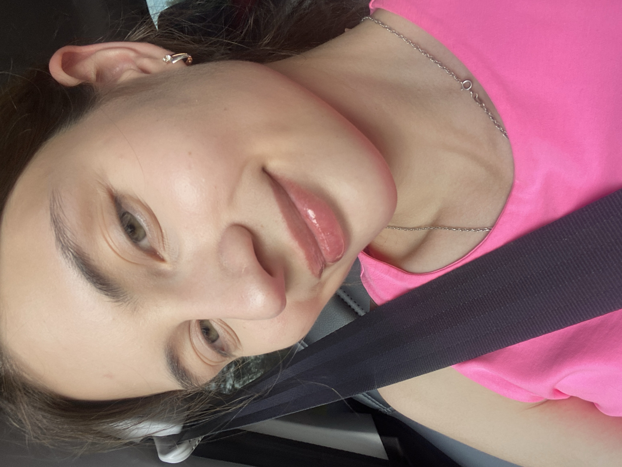 A young woman with light skin and brown hair is captured in a close-up, forward-facing selfie. She is wearing a bright pink tank top and a silver chain necklace. A black seatbelt is diagonally across her chest. Her eyes are a light green color and her lips are glossed in a peachy-pink hue. She is smiling faintly, with her head tilted slightly to her right. Her left ear is visible, adorned with a small hoop earring. The background is dark and out of focus, suggesting an indoor setting, possibly a car. The image is well-lit, highlighting the woman's features. There are no discernible elements in the image that would indicate a specific location within the city of Kamsskoye, Russia.