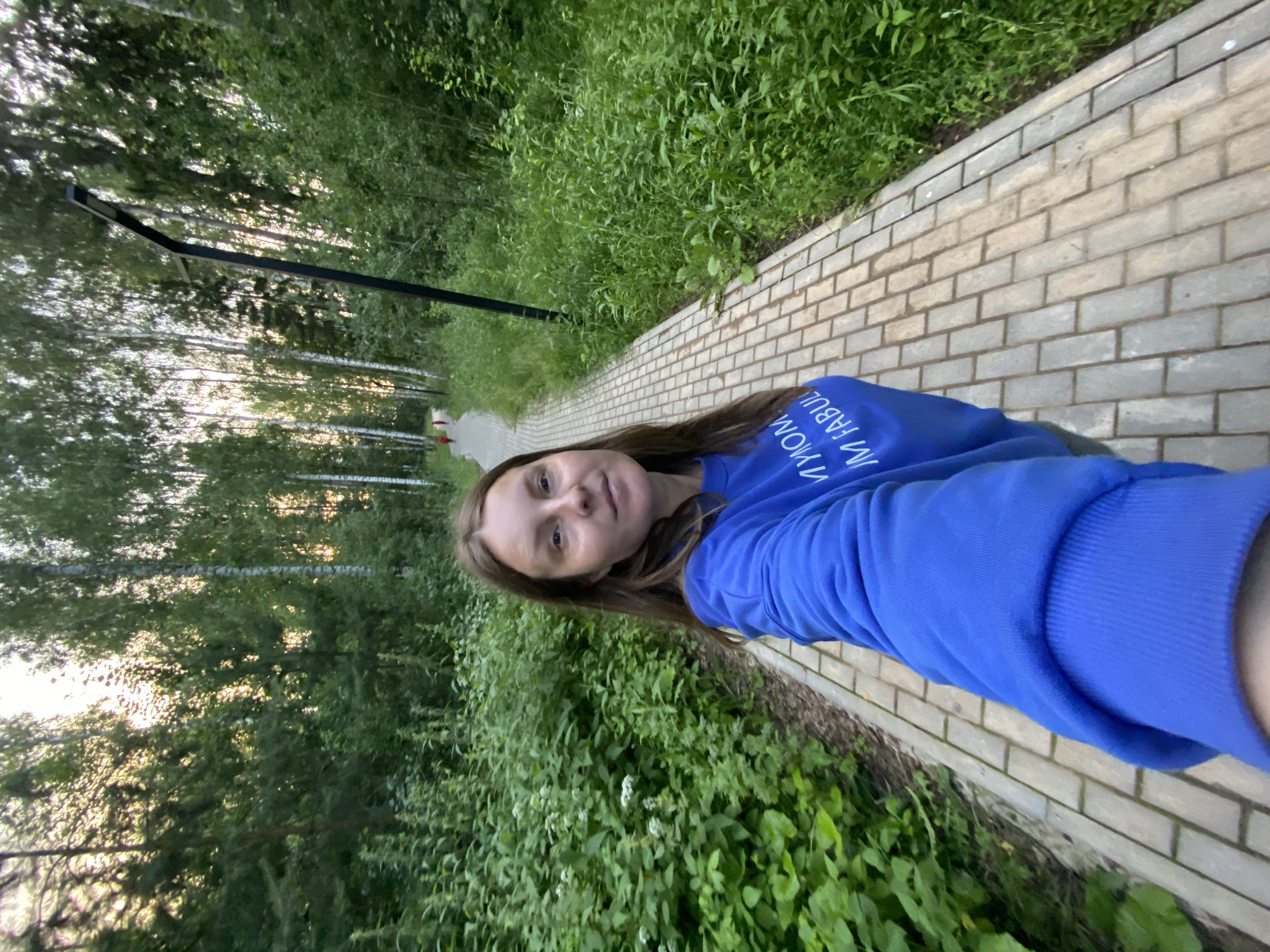 A young woman with long brown hair is taking a selfie while walking on a brick pathway through a wooded area. She is wearing a bright blue sweatshirt with white text on the sleeve that reads "I'M FABUL". The sun is setting, casting a warm glow through the trees. The greenery is lush and abundant, suggesting a warm season. The overall mood appears relaxed and content. The pathway is lined with trees on one side and dense vegetation on the other. A black lamppost is visible in the background. The image is oriented vertically.