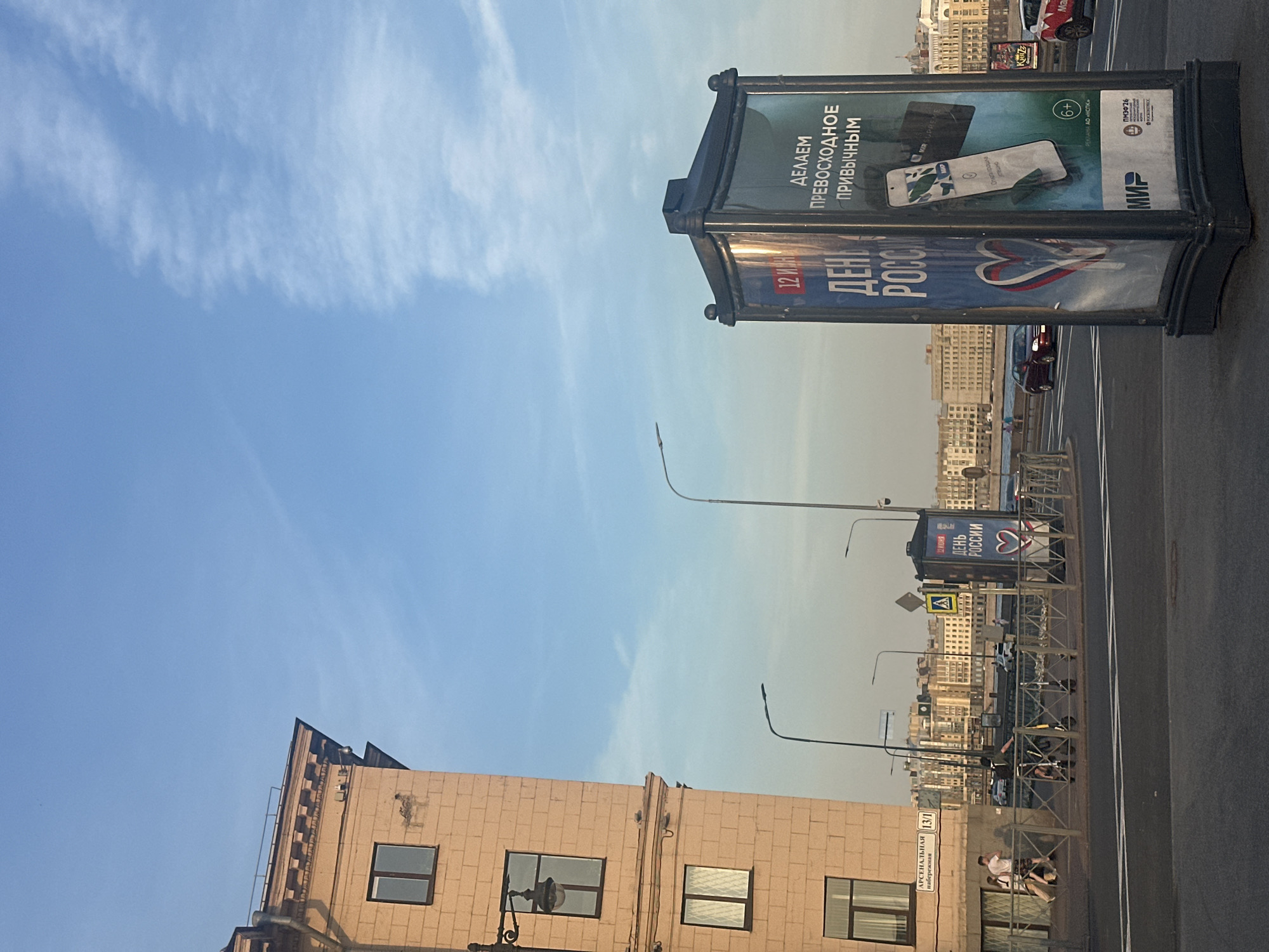 The image shows a street scene in a city with a clear blue sky and some wispy clouds. On the left is a light-colored, multi-story building with several windows. In the foreground, a modern advertising pillar is visible, displaying posters with Russian text and images, including a smartphone and a heart graphic. The pillar is located on a sidewalk next to a road with traffic. Several streetlights and a pedestrian fence are also present. In the background, there are more buildings, a body of water, and a bridge. A few people and a scooter are on the sidewalk near the building. Cars are visible on the road. The overall impression is a sunny day in an urban environment.