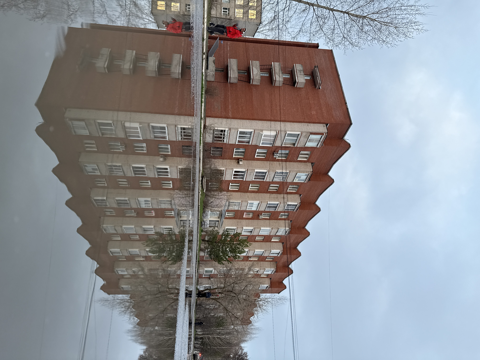 The image captures an urban or suburban scene featuring a multi-story building prominently reflected in a large, still body of water, likely a puddle on a wet surface. The building, constructed with red-brown brick on its upper sections and lighter gray/beige sections with numerous white-framed windows on lower floors, exhibits a distinctive stepped roofline. Several external structures, possibly air conditioning units or decorative elements, are visible on its facade.
A person wearing a bright red jacket, dark pants, and a dark hat stands near the edge of the reflective surface, close to the building. Another person is partially visible further away in the reflection. The still water creates an almost perfect, inverted reflection of the building, trees, and sky, dominating the foreground and lower portion of the image.
Bare trees, indicative of late fall, winter, or early spring, line the area behind the puddle and extend into the sky. The sky is overcast, suggesting a cloudy day. Overhead wires are faintly visible against the sky. Another, less distinct building can be seen in the background. The reflective quality of the water is a key element, blurring the line between the real structures and their mirror images. No visible text is present.