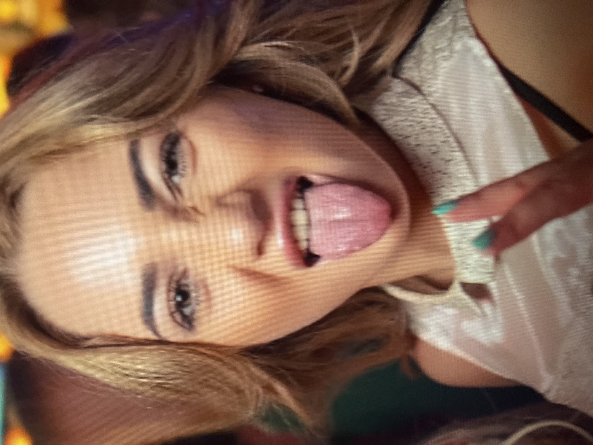 A medium shot captures a young Caucasian woman with blonde hair and light eyes sticking her tongue out and making a peace sign. She is wearing a light-colored, possibly satin, top with a decorative collar. Her fingernails are painted a teal color. The background is blurred, suggesting an indoor, possibly dimly lit setting like a club or party, with hints of other people and colored lights. There is no discernible location within the city administration of Qarağandı city, Kazakhstan.