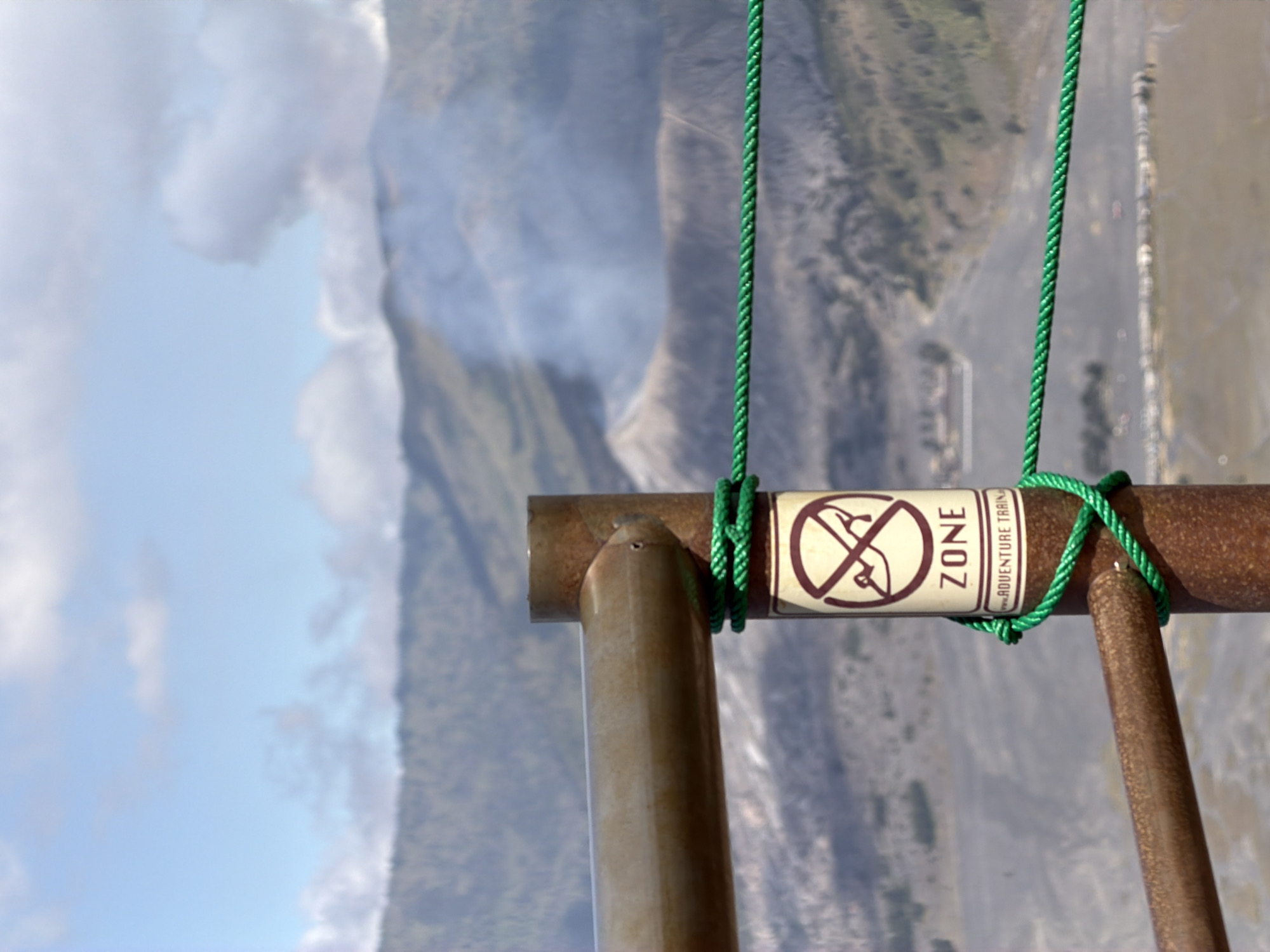 The image depicts a close-up view of a metal fence post with a sign attached, in the vicinity of Mount Bromo, Cemorolawang, Indonesia. The sign reads "ZONE" and has a red circle with a diagonal line across it, superimposed on an image of a person wearing hiking boots, indicating a restricted area. Below "ZONE" is smaller text that appears to say "ADVENTURE TRAIL". A green rope is tied around the fence post, securing a section of the fence. In the background, a hazy, mountainous landscape is visible, with a plume of smoke rising from what appears to be a volcanic crater. The sky is partly cloudy, with blue sky visible between the clouds. The scene suggests a volcanic observation point or a trail leading towards a volcanic area. There are no people visible in the immediate foreground.