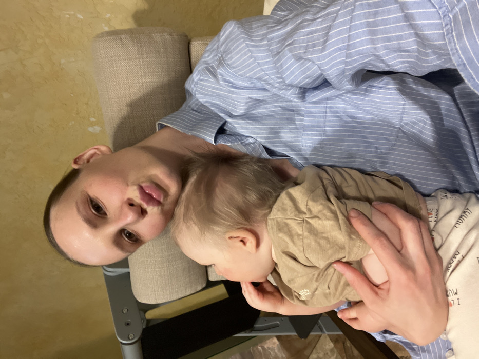 The image captures a tender moment between a woman and a baby in an indoor setting. The photograph is oriented with the subjects turned 90 degrees to the left, appearing to be lying down or leaning back.
A woman, likely an adult, is positioned on the left side of the frame, looking directly towards the camera. She has light skin, dark eyes, and her hair appears to be pulled back. Her lips are slightly parted, and she has a calm, relaxed expression. She is wearing a light blue button-down shirt with thin white vertical stripes.
She is holding a baby or young toddler close to her, with the baby's head resting on her chest or shoulder, facing away from the camera. The baby's face is mostly hidden, but light-colored hair is visible. The baby is dressed in a light brown or tan short-sleeved top over a lighter-colored long-sleeved onesie or shirt. The woman's right hand is gently supporting the baby's back and side. A small part of the baby's hand is visible near its mouth or chin.
In the background, a textured wall with a warm, yellowish-golden hue is visible. To the left of the woman's head, there is a cushioned, upholstered backrest or armrest, possibly part of a high chair, sofa, or chair, in a neutral, light beige fabric. Below the baby and the woman's hand, parts of what appear to be the structure of a high chair or piece of furniture are visible.
Notable text is visible on the baby's under-layer clothing. Repeated words such as "MUMMY" and "DADDY" are printed in a pattern, and a partial phrase "I LOVE MUMMY" can also be deciphered near the bottom right corner of the frame. The overall atmosphere of the image is peaceful and intimate, suggesting a moment of comfort or rest between the mother and child.