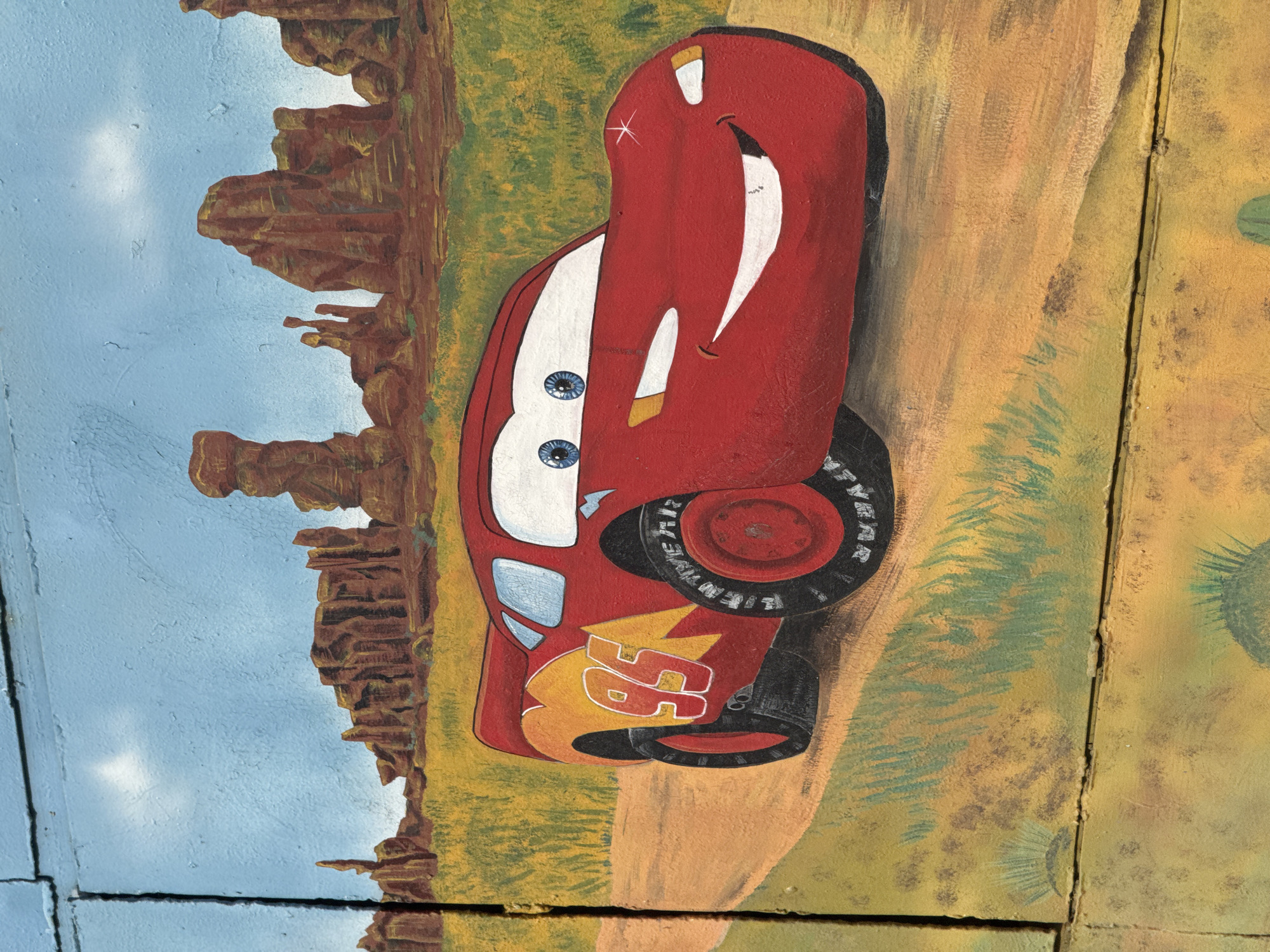 A painted mural, likely on a textured wooden surface exhibiting vertical and horizontal seams and minor paint chipping, depicts Lightning McQueen from Disney/Pixar's *Cars* franchise in a desert landscape. This artwork is located in Chaikovsky, Russia.
The central subject is the anthropomorphic red race car, Lightning McQueen, positioned on a light brown dirt road and angled slightly towards the viewer's right, facing forward. The car exhibits large blue eyes, a broad white smile, and yellow headlights. Its side features a prominent yellow lightning bolt graphic with the white number "95" outlined in red. The tires are black with visible tread patterns.
The background consists of a sparsely vegetated desert environment. Light green and yellow brush-like flora are scattered across sandy, ochre-colored ground. Numerous reddish-brown rock formations, characteristic of buttes or mesas, rise in the mid-ground and background under a light blue sky containing minimal, indistinct white cloud formations.