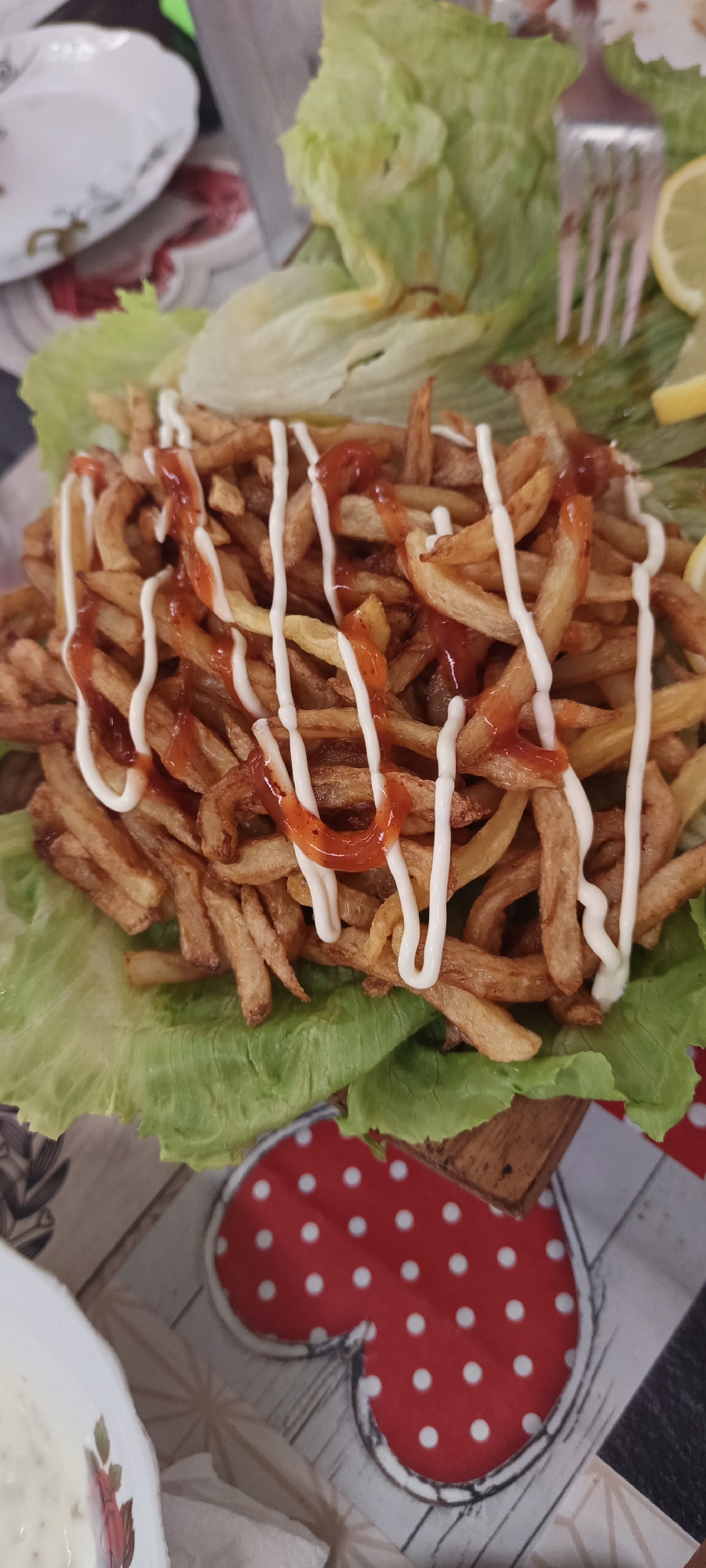 A close-up, overhead shot shows a plate of french fries topped with ketchup and mayonnaise. The fries are served on a bed of lettuce, with a lemon slice and a fork visible in the background. The plate is on a wooden table with a red and white polka dot placemat. The image appears to have been taken in Khudat, Azerbaijan, a city known for its culinary scene.