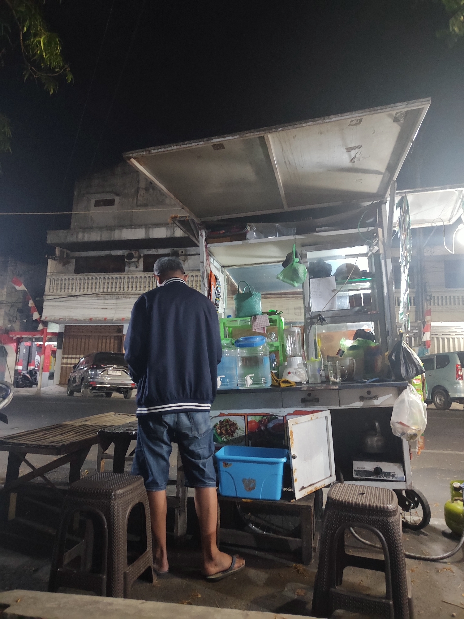 A man stands facing away from the camera, wearing a dark blue jacket with white trim and denim shorts. He is positioned behind a mobile food cart, which is set up on a street at night. The cart has a white canopy and is stocked with various items including large water dispensers, a blender, cups, and what appears to be a small stove inside a cabinet. In the background, there is a multi-story building with balconies and a parked silver car. Other vehicles and a propane tank are also visible in the scene. The lighting suggests it is evening or nighttime.