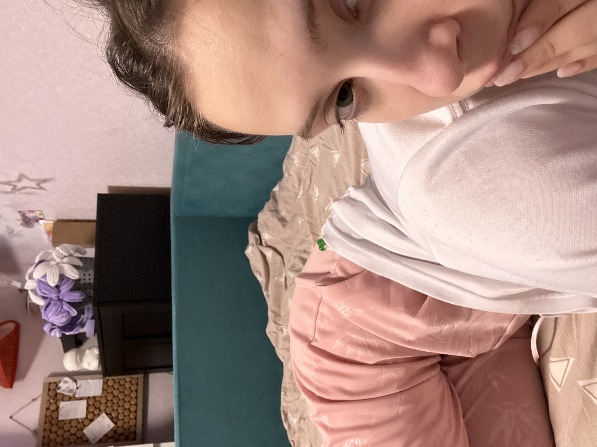 A rotated close-up shot captures a person's face with brown hair and fair skin. They are wearing a white sweatshirt over a pink, silky top or robe. Their hand is near their mouth, possibly touching the lips. The background consists of a turquoise headboard, beige patterned bedding, and a collection of objects including purple and white flowers, a black rectangular object, a red item, and a tray of round, brown objects with small white pieces on top. There is also a piece of paper and a star on the wall. The image does not contain enough information to verify the location in the city of Прикамский, Russia.