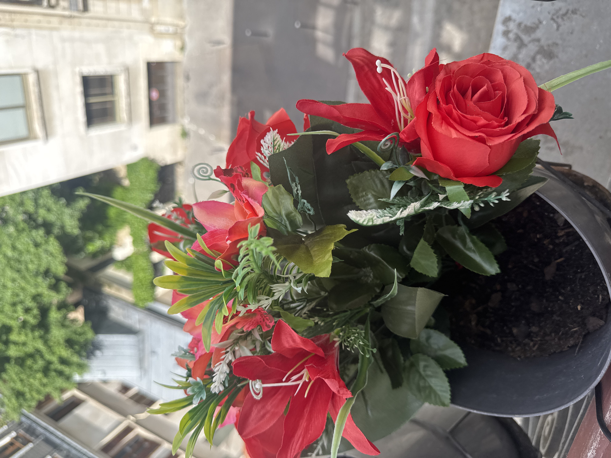 The image shows a close-up of a pot of artificial red flowers (roses and lilies) on a balcony, with an urban view of buildings in the background. The buildings suggest an older architectural style. The presence of vegetation and landscaping elements in the distant buildings may indicate residential settings or courtyards. Given these attributes, the location is possibly Paris, France.