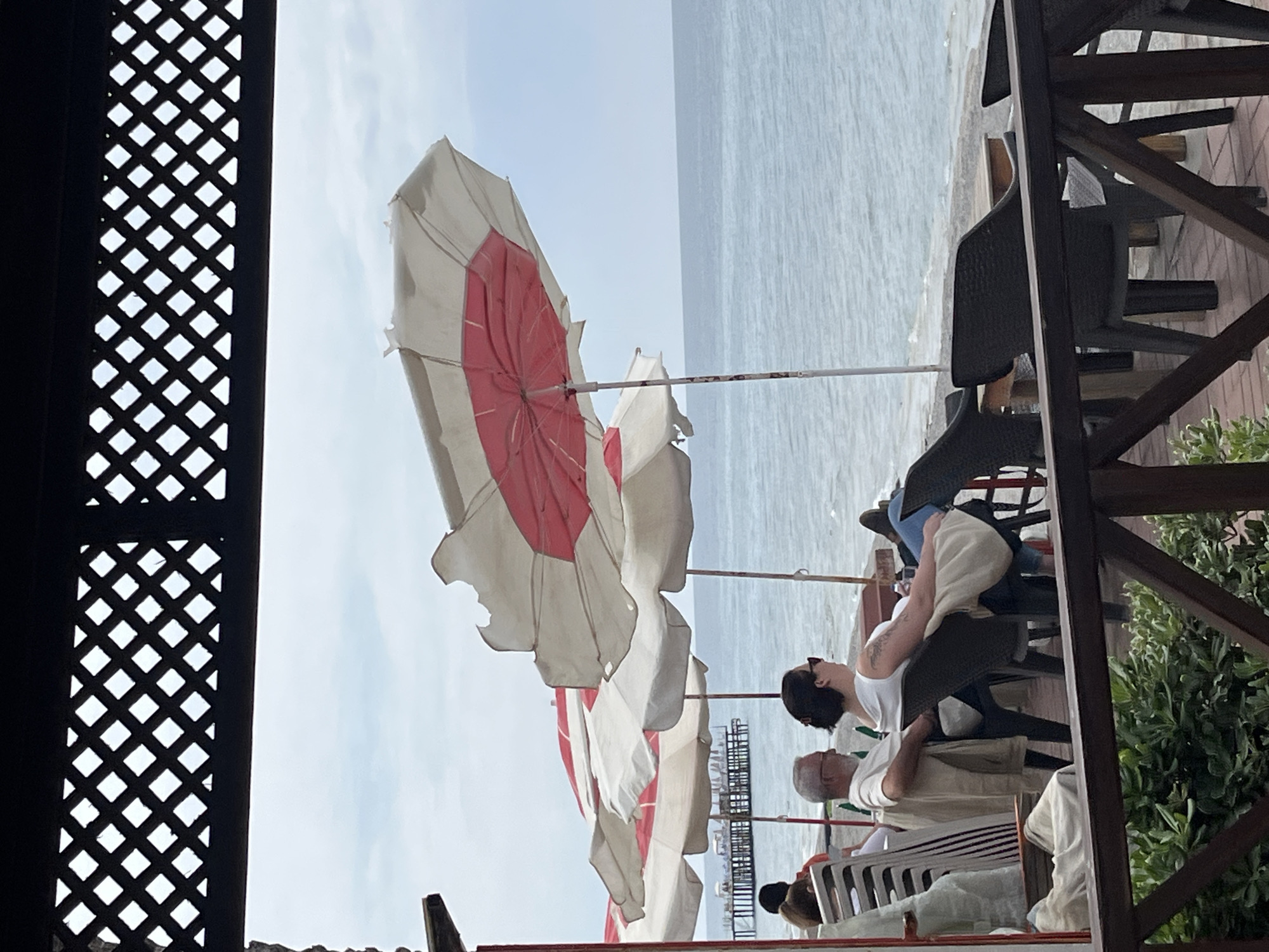 The image, taken in Alanya, Turkey, shows an outdoor cafe or restaurant setting. In the background is a view of the sea under a clear, overcast sky. Several beach umbrellas with white and red patterns are partially visible. A partially obscured wooden lattice is in the left foreground. Adjacent to the sea is a pier. Seated at the tables are several people, one of whom is wearing a white tank top, and another a white shirt. Additional chairs are stacked nearby. The right side of the frame contains a wooden structure and greenery.