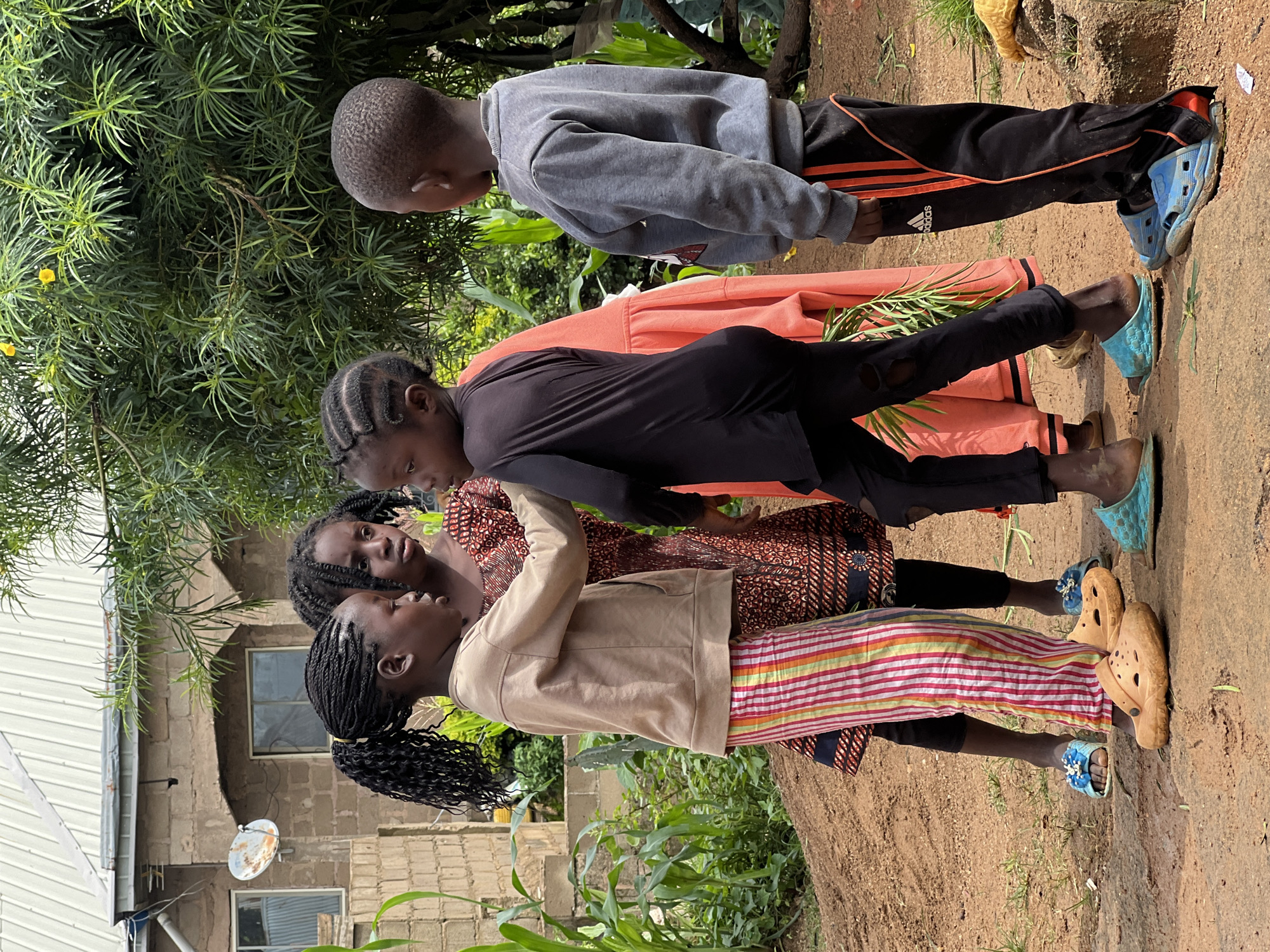 In the city of Jos, Nigeria, four children are gathered in an outdoor setting. A young girl with her hair in braids and a high ponytail wears a tan sweatshirt and colorful striped pants, with tan clogs on her feet. Beside her, another girl with braided hair wears a patterned orange and brown dress. A third child, facing away from the camera, wears a black long-sleeved shirt and black pants with orange stripes, and turquoise slides. A fourth child, also facing away, wears a grey sweatshirt and black pants with orange stripes, and blue slides. They appear to be interacting with each other. The environment includes a building with a metal roof in the background, lush green foliage, and a dirt ground.