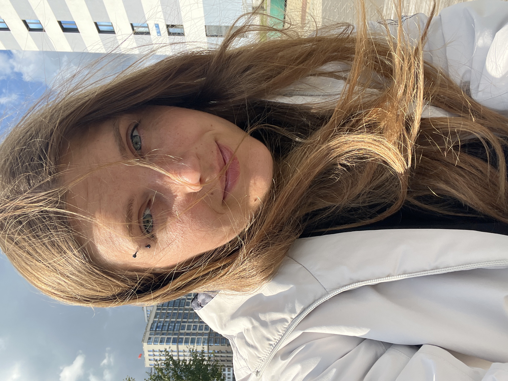 This is a selfie of a young woman with long, blonde hair and blue eyes, taken outdoors in the city of Minsk, Belarus. She is wearing a light-colored jacket over a dark shirt. A small eyebrow piercing is visible on her left eyebrow. In the background, modern apartment buildings are visible, one of which is a white building with a grid pattern of dark windows. The sky is partly cloudy, and the sun is casting shadows. The overall impression is a candid shot taken on a bright, windy day.